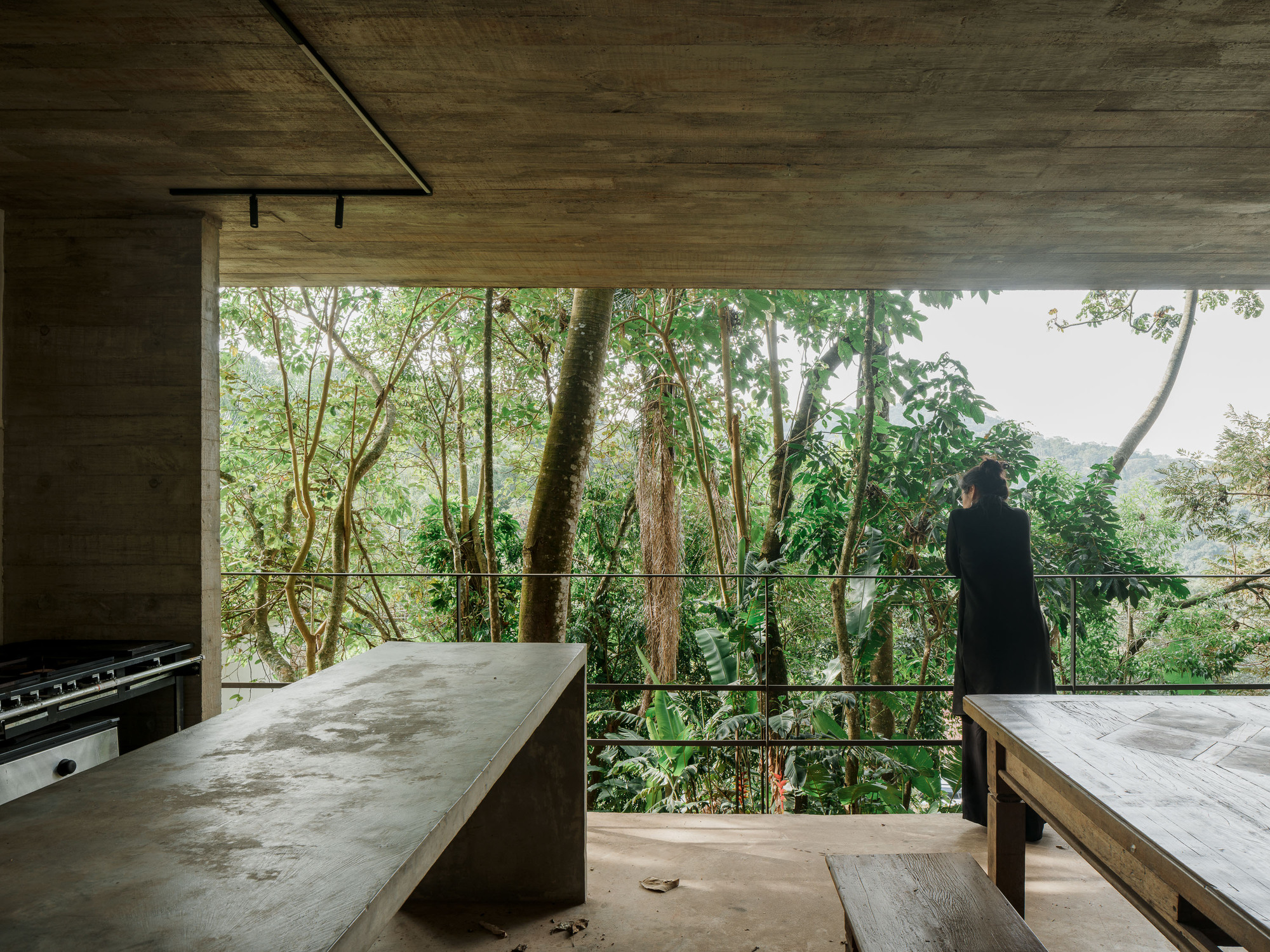 Open kitchen with concrete counters and timber ceiling looking out to forested hillside through glass railings
