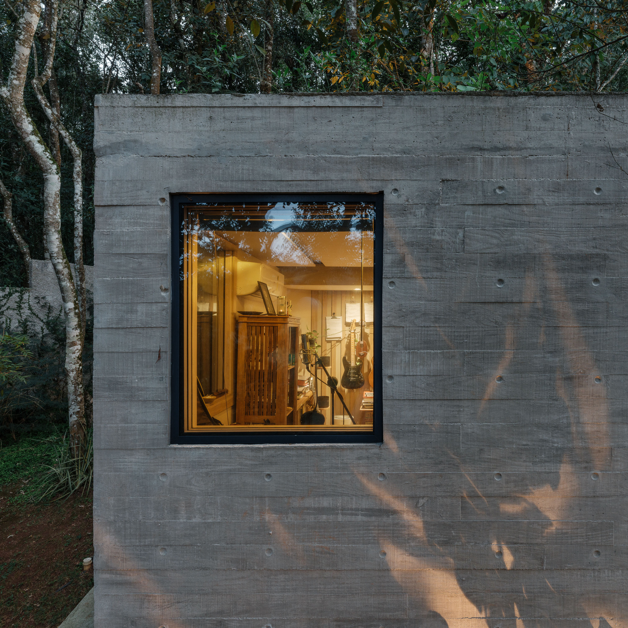 Single window cut into board-formed concrete wall illuminated at dusk with tree shadows across the surface