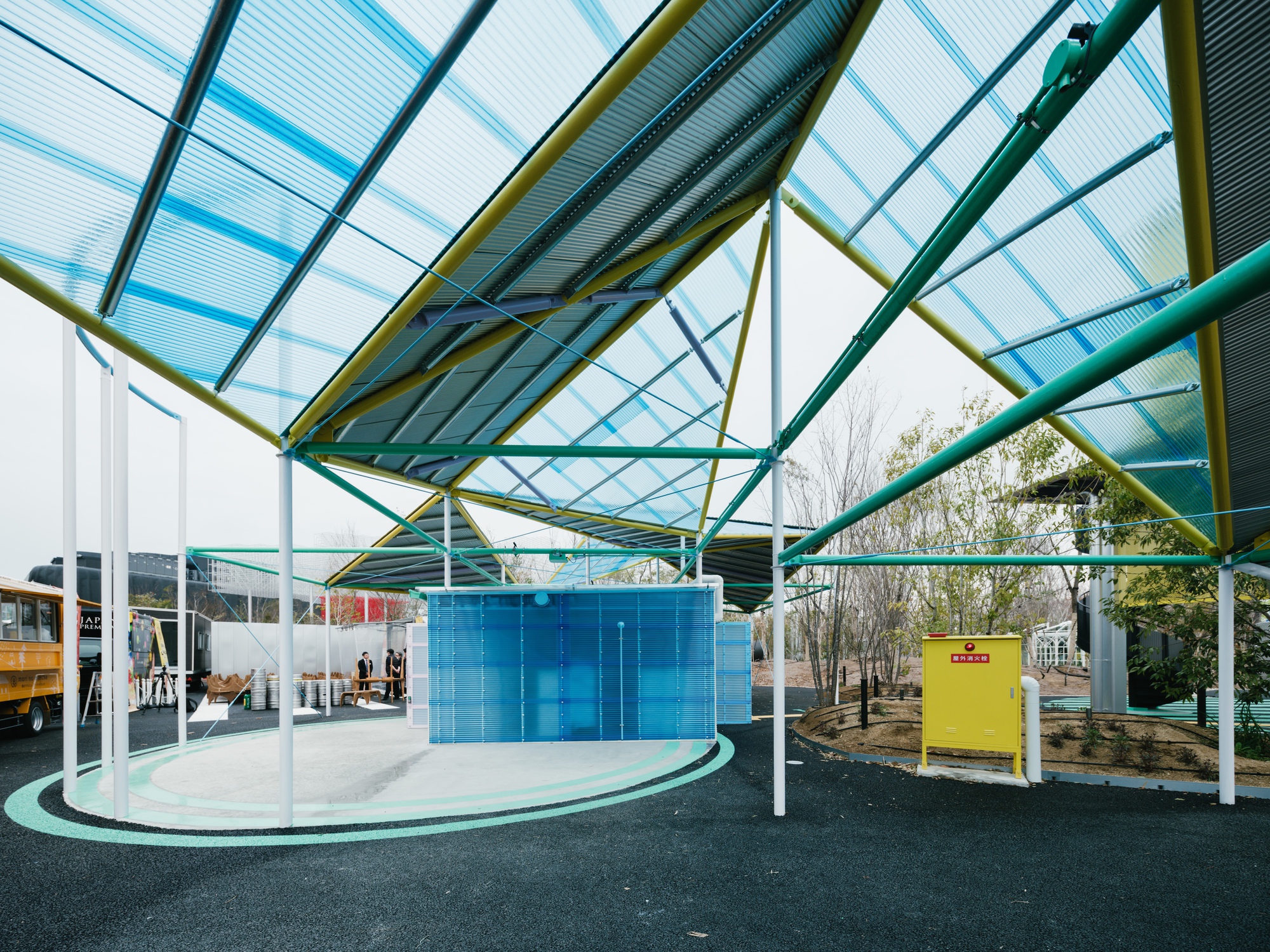 Covered outdoor plaza with colored steel columns and translucent corrugated canopy over asphalt ground with painted graphics