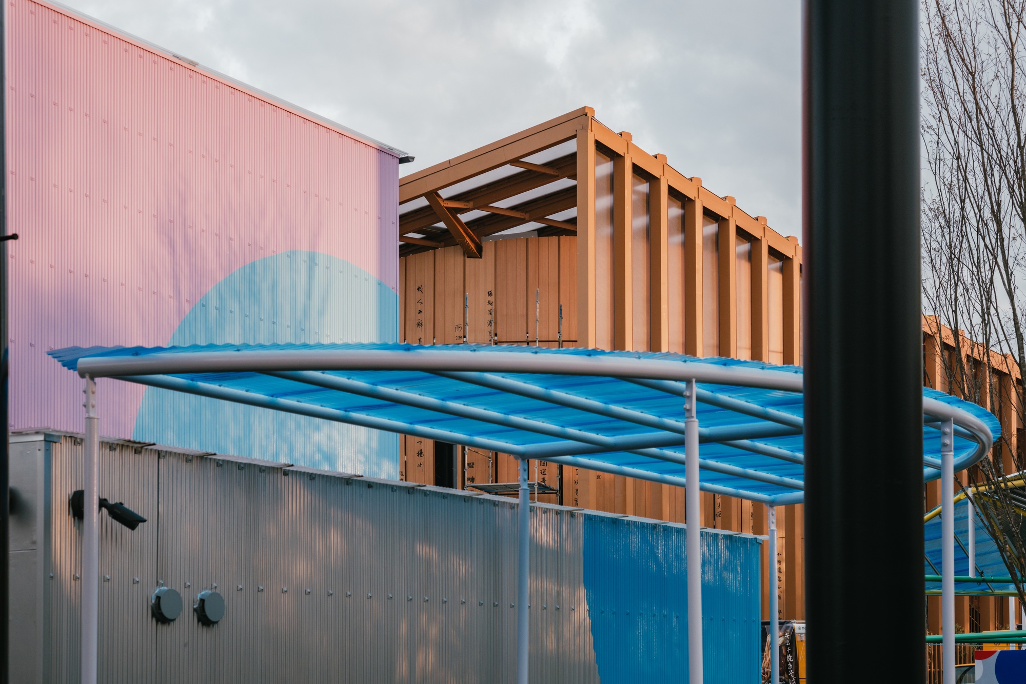 Corrugated metal facade with blue painted circle detail beneath timber framed roof structure and tubular awning