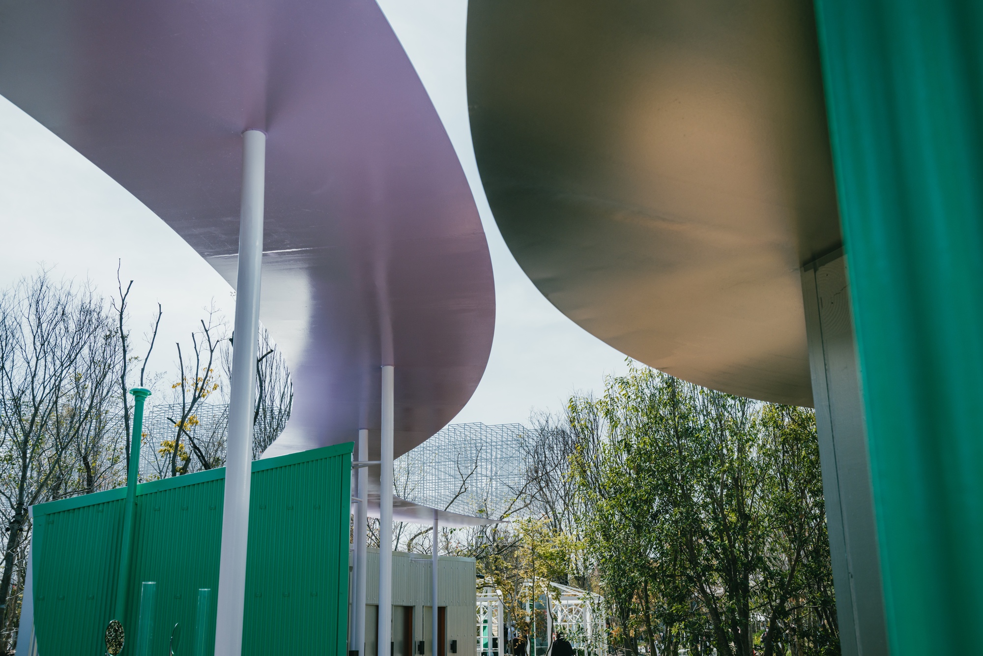 Underside of curved colored canopy panels supported by white cylindrical columns with green corrugated wall beyond