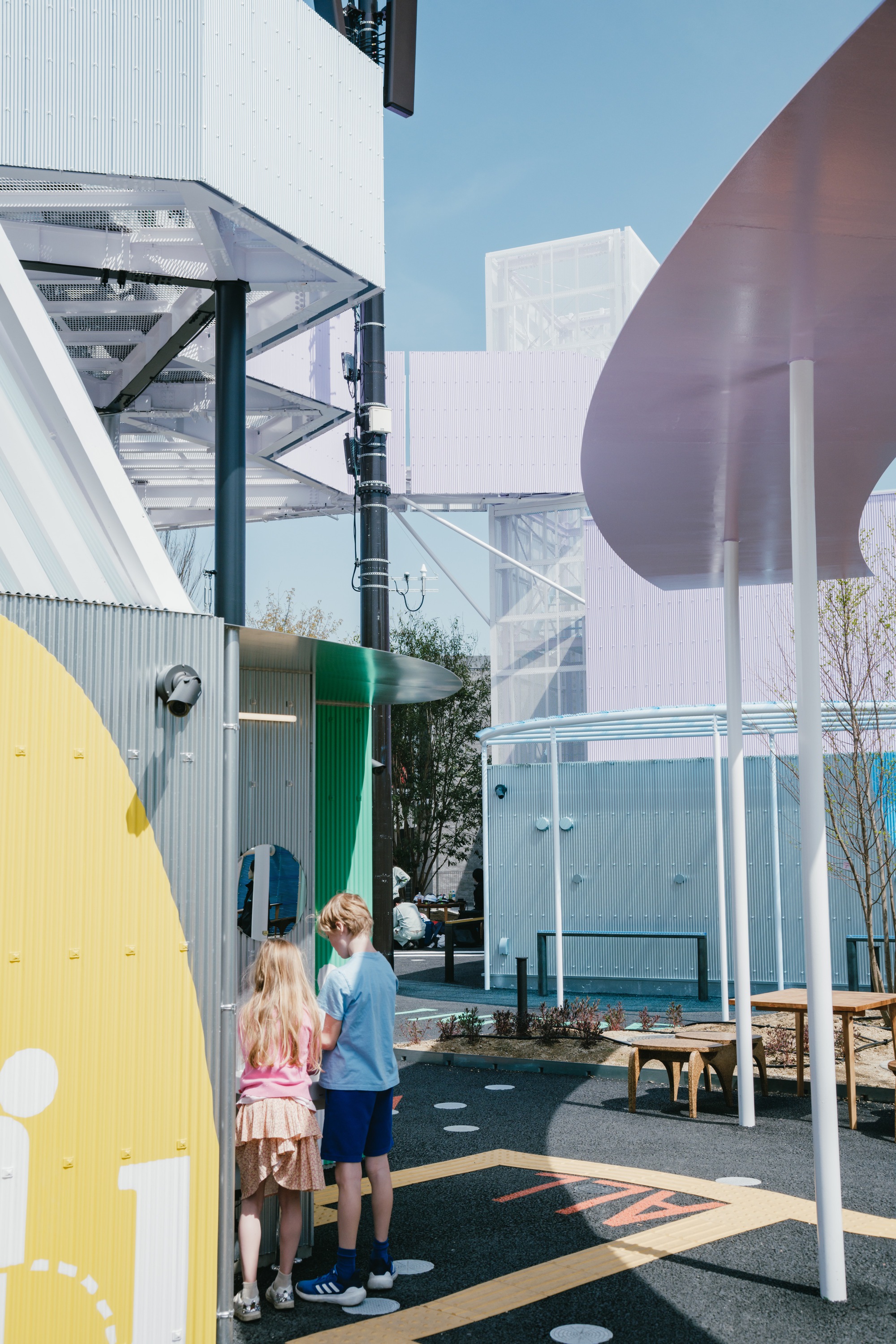 Outdoor courtyard with corrugated metal volumes, coloured panels, and two children looking up at a canopy structure