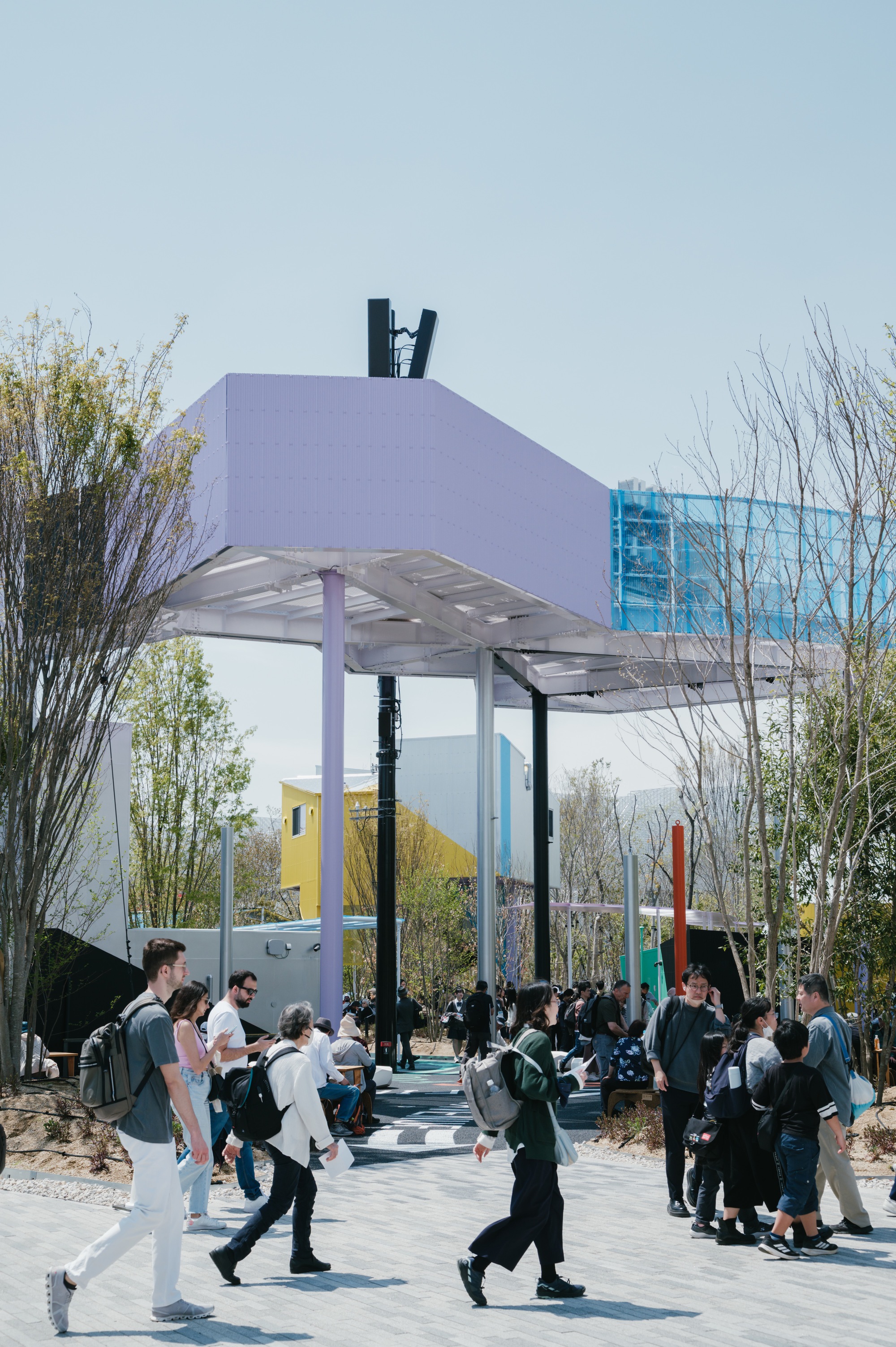 Canopy structure with steel columns and perforated metal deck sheltering a pathway filled with visitors