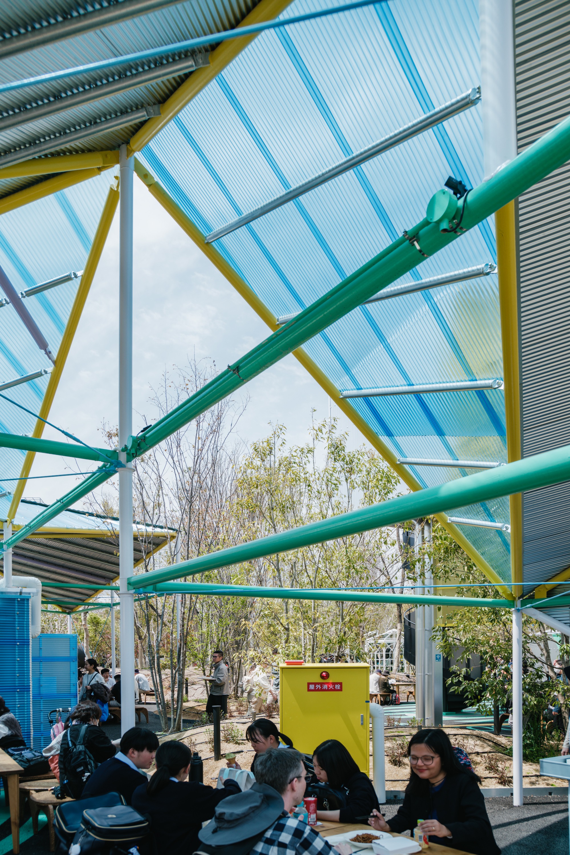 Underside of the canopy showing yellow and green painted steel trusses with translucent corrugated roofing panels