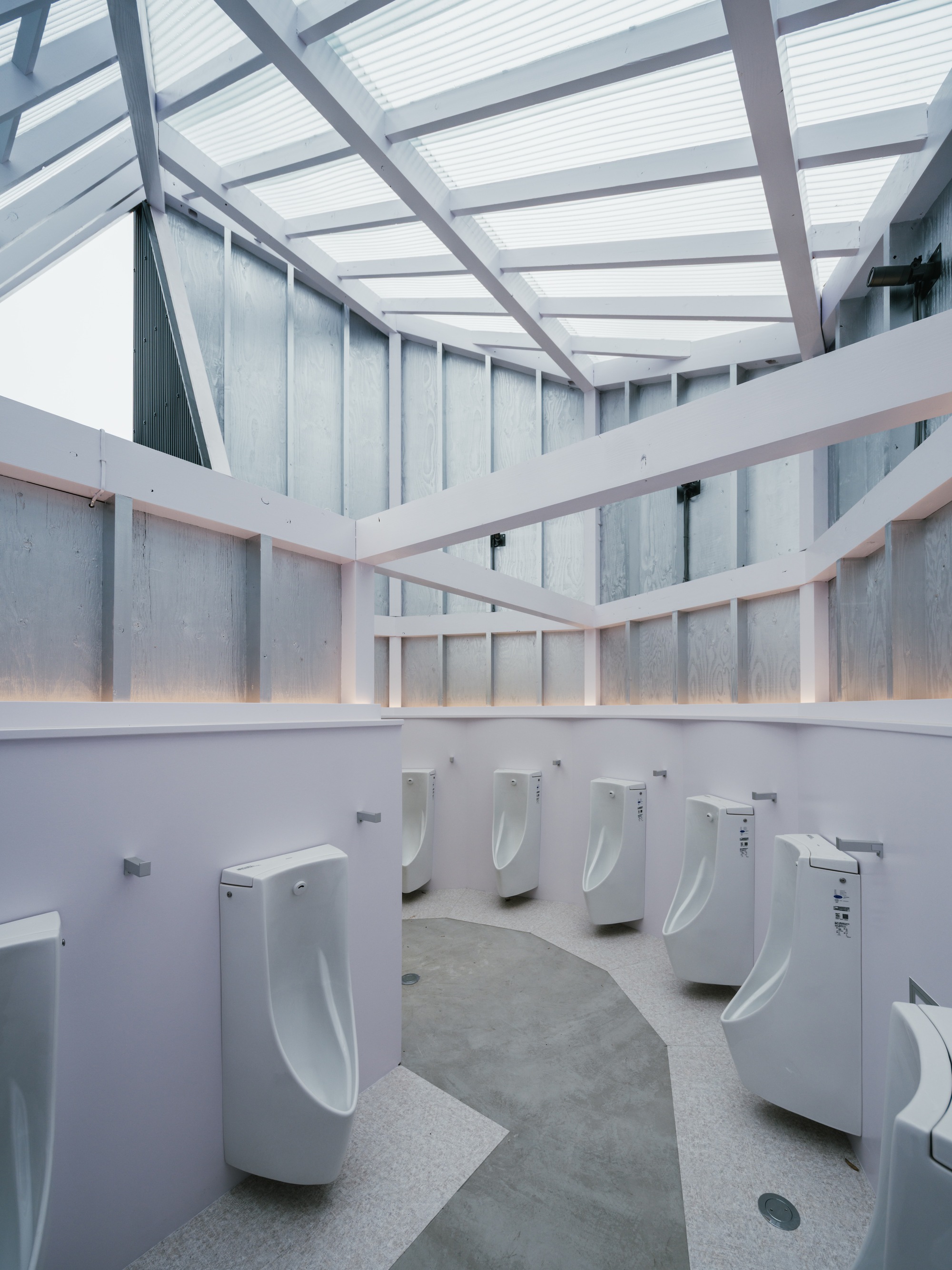 Circular restroom interior with row of white urinals beneath a translucent polycarbonate skylight and metal framing