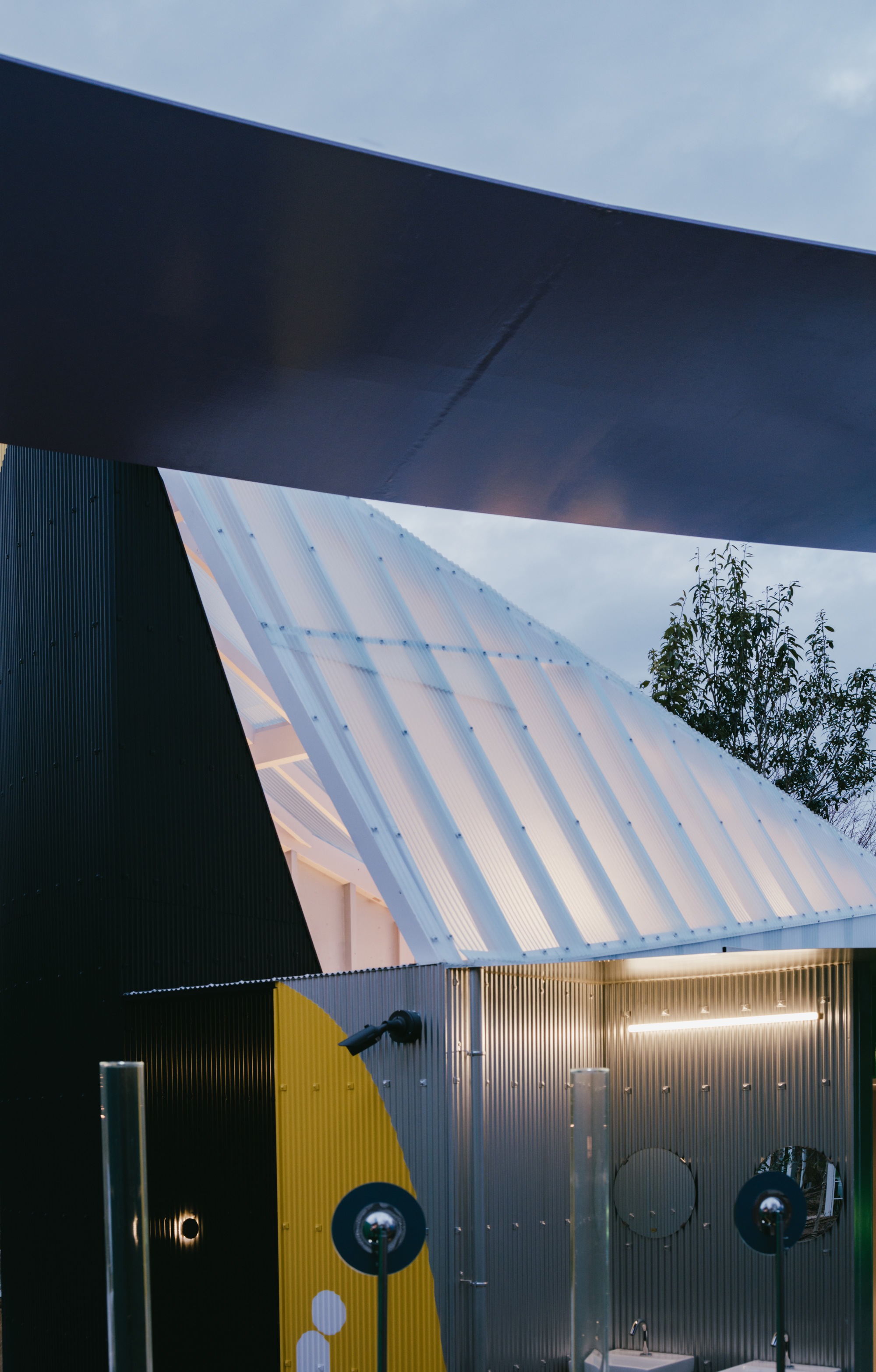 Angled corrugated facade and dark overhang meeting at twilight with bare trees visible