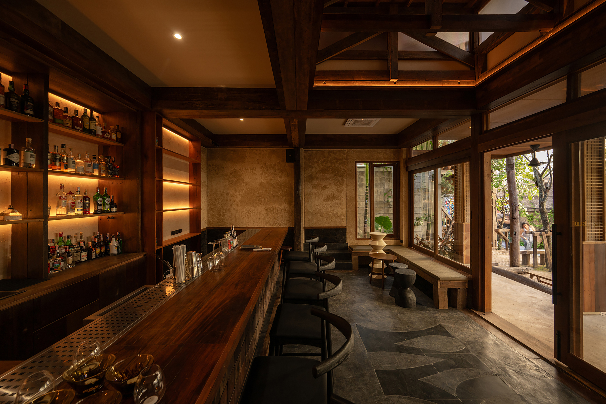 Interior bar with backlit shelving, timber post-and-beam structure and glazed doors overlooking a planted garden