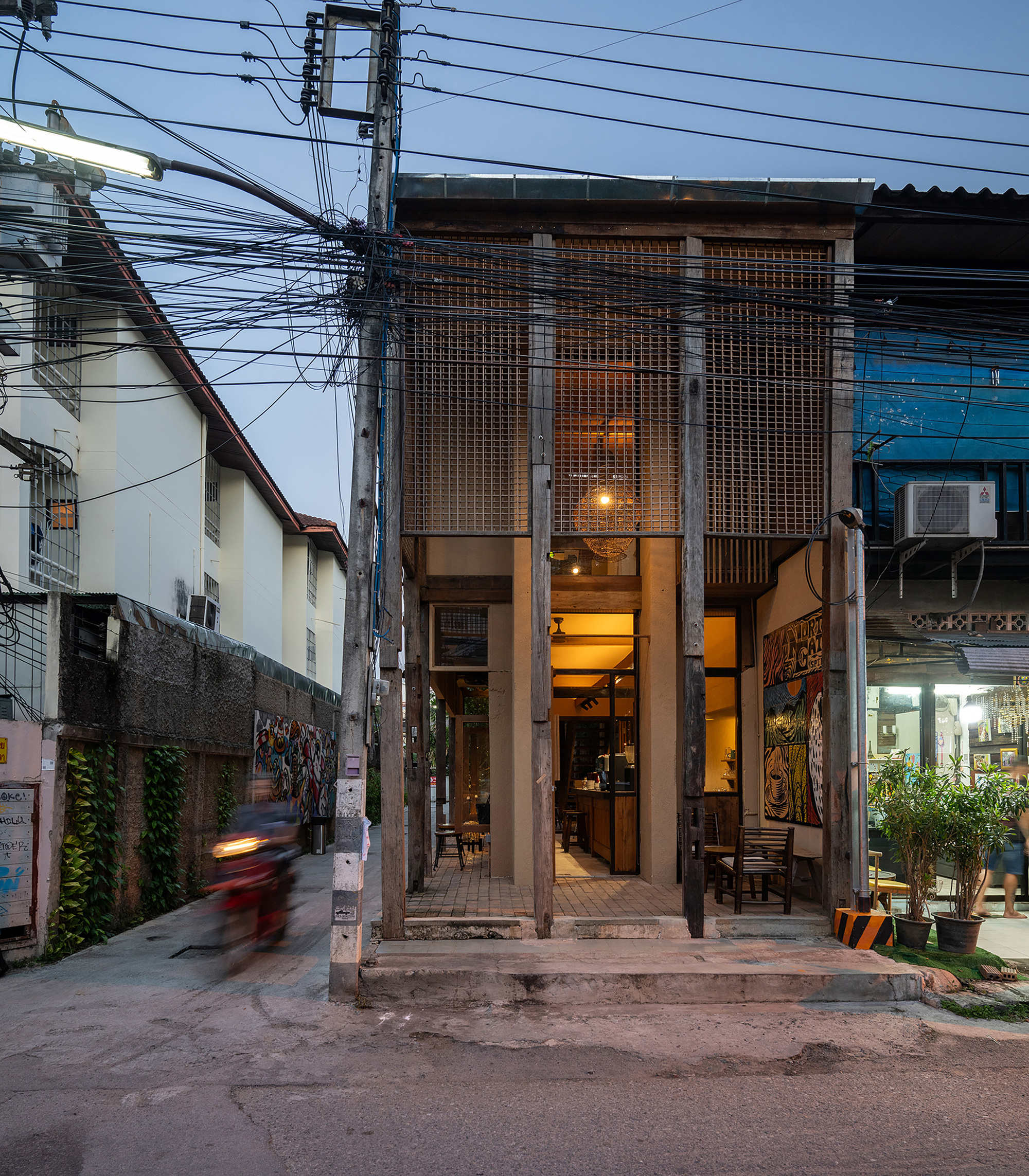 Narrow street facade with perforated metal screen panels, exposed columns and overhead power lines at twilight