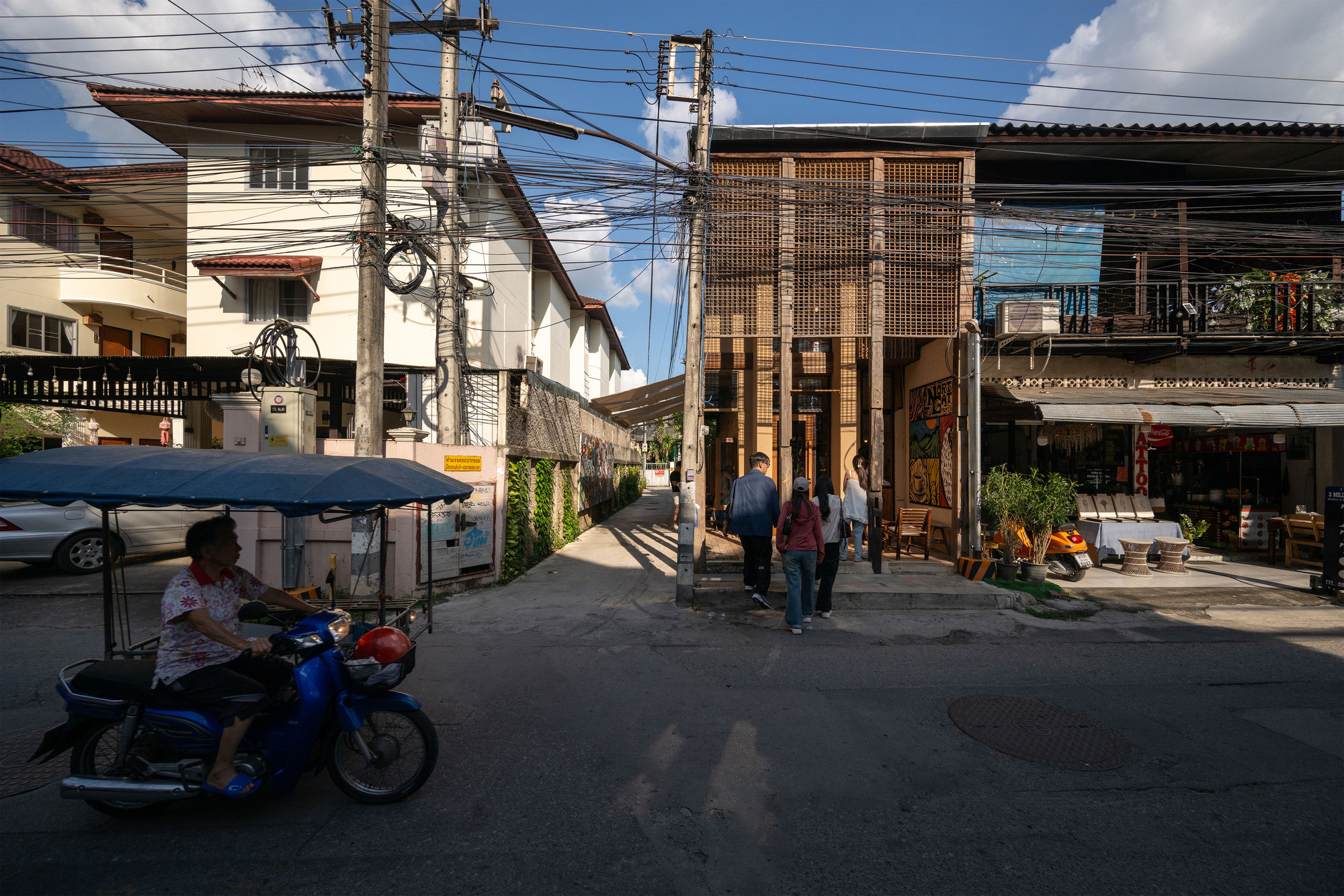 Street view of a metal screen facade with pedestrians and a motorcyclist passing beneath overhead wires