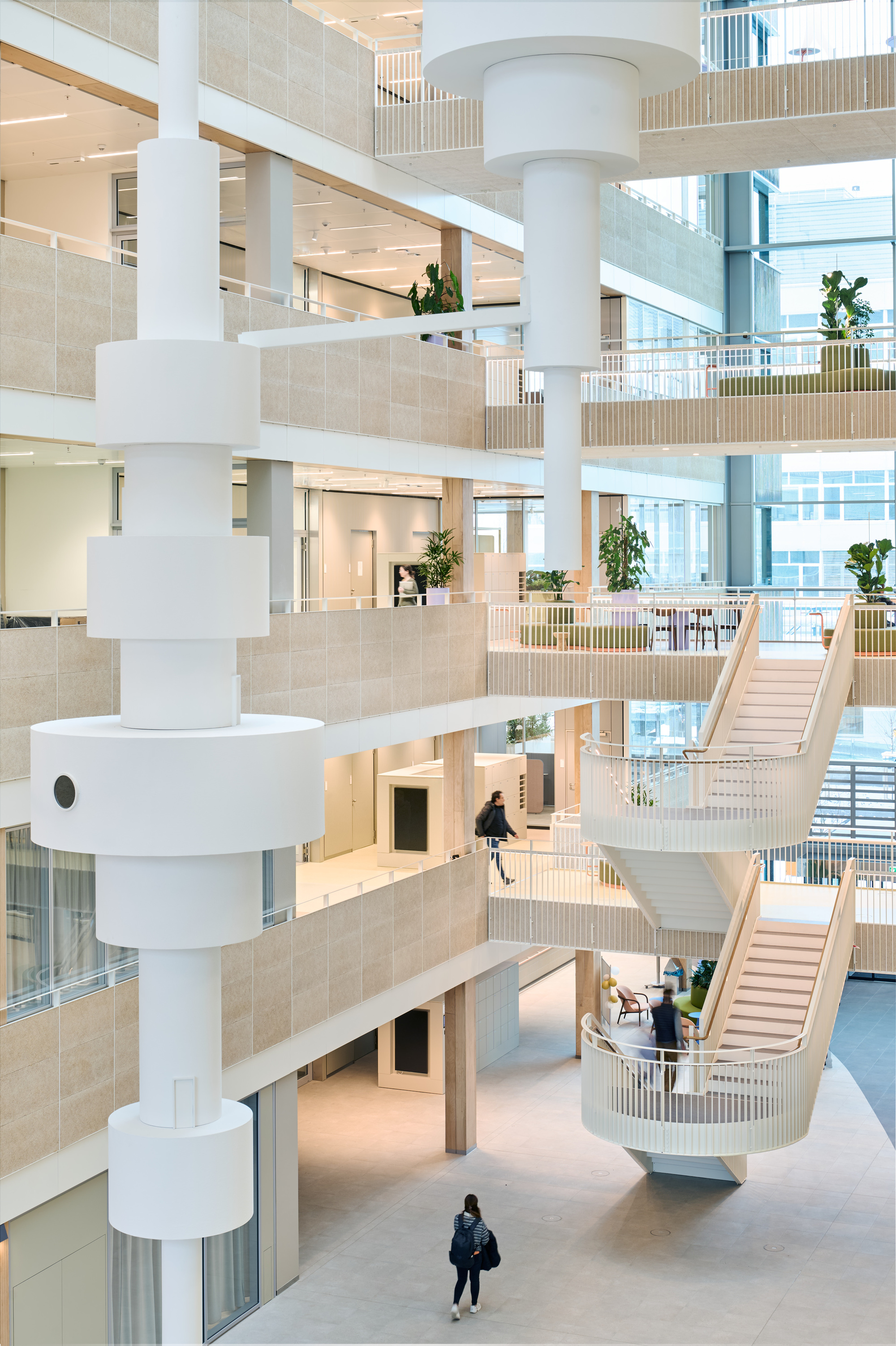 Multi-storey atrium with white cylindrical columns, timber balustrades and two pedestrians crossing the polished floor