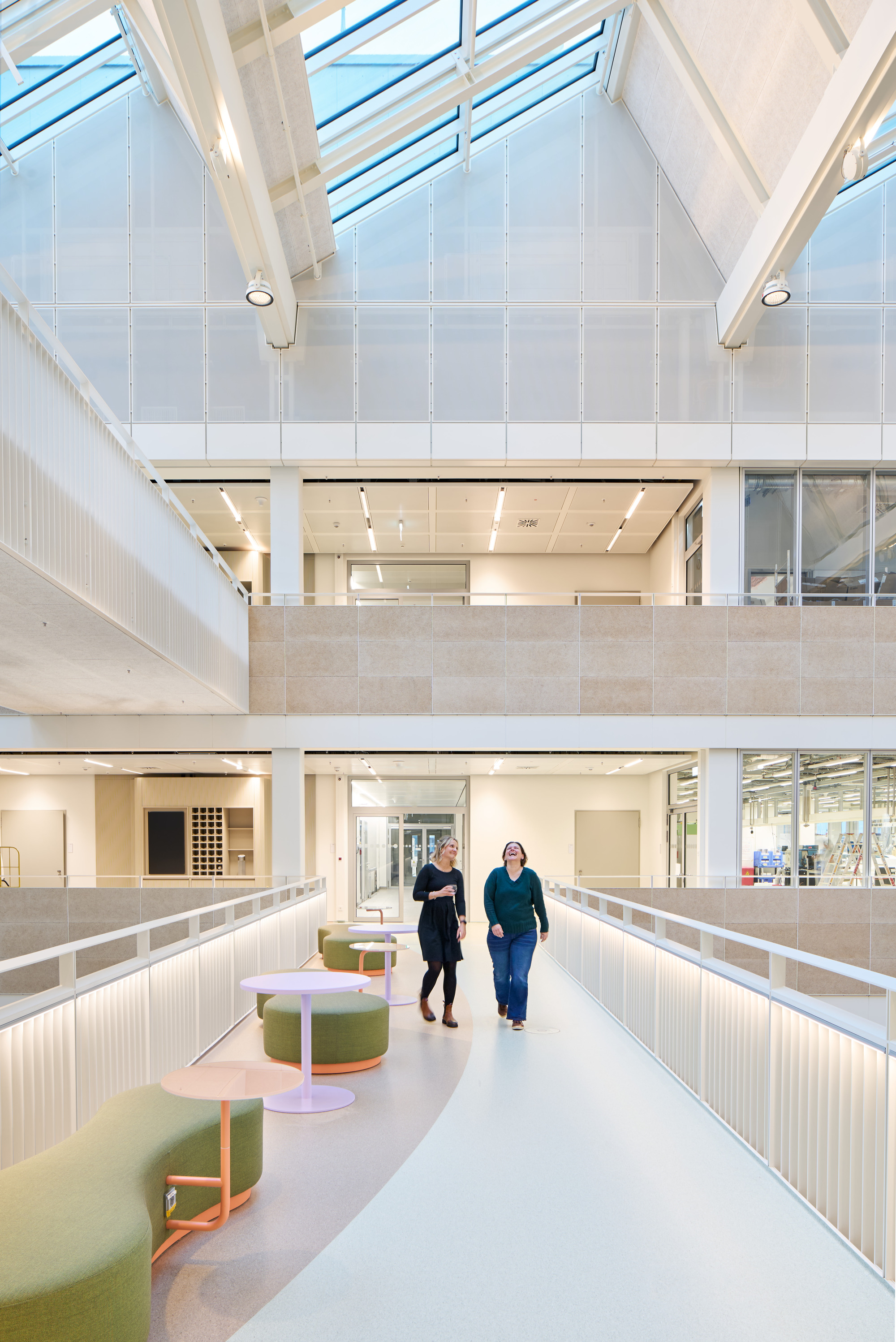 Upper corridor with green upholstered seating and two people walking beneath a skylit ceiling