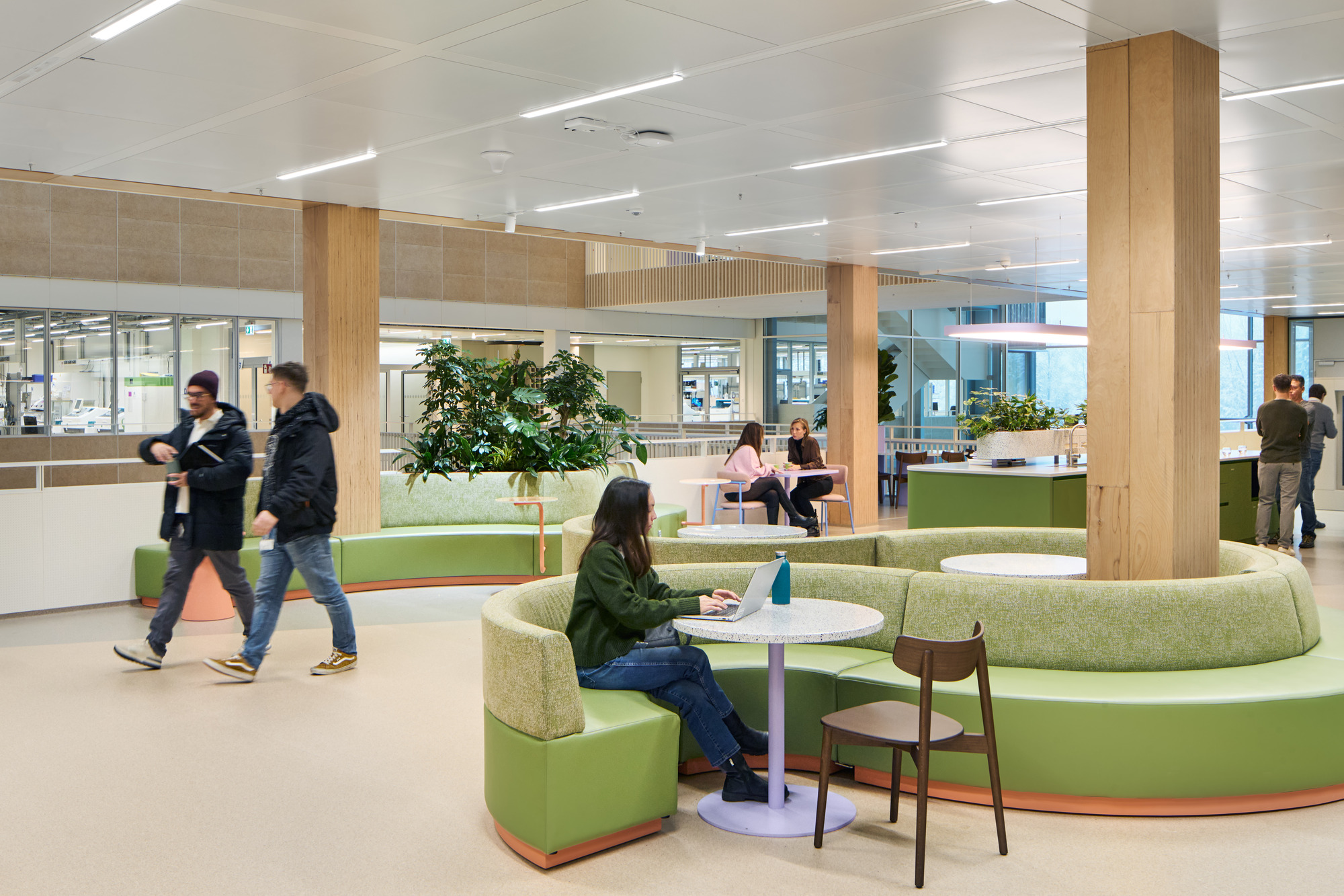 Open office lounge with curved green banquette seating, timber columns and potted plants among casual workers