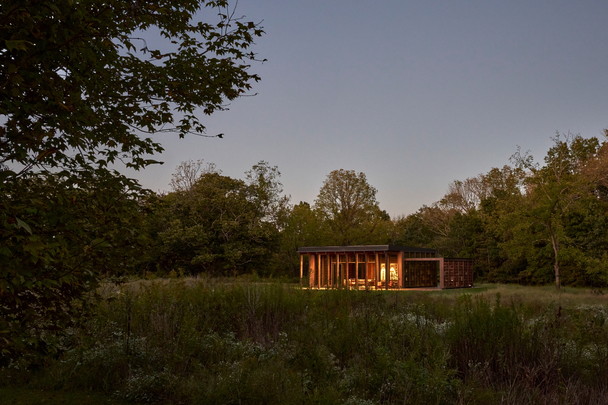 Distant view of the glazed pavilion glowing at dusk across a meadow with tall grasses and scattered trees