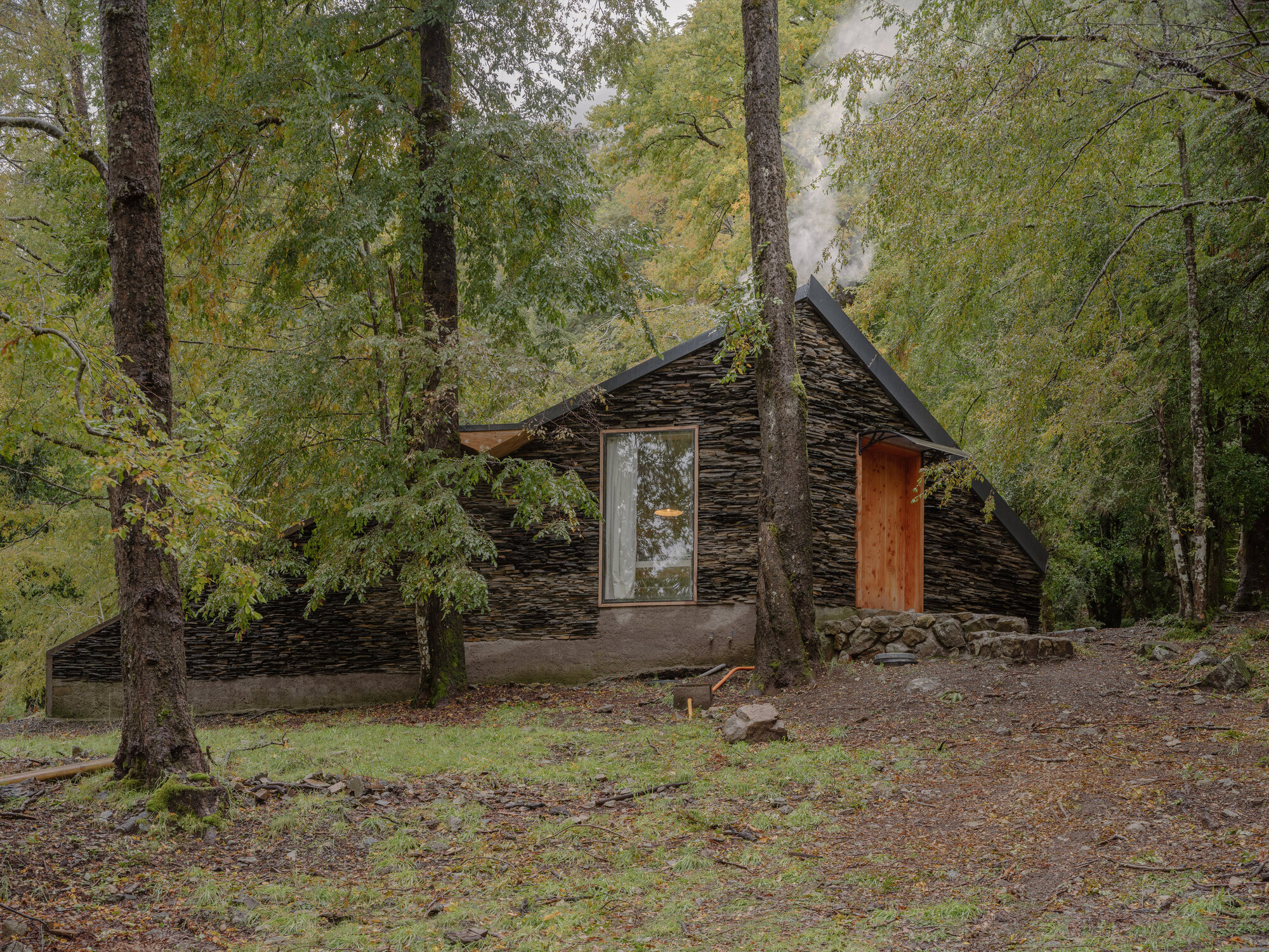 Angled view of the layered stone roof with timber door and chimney smoke among forest trees