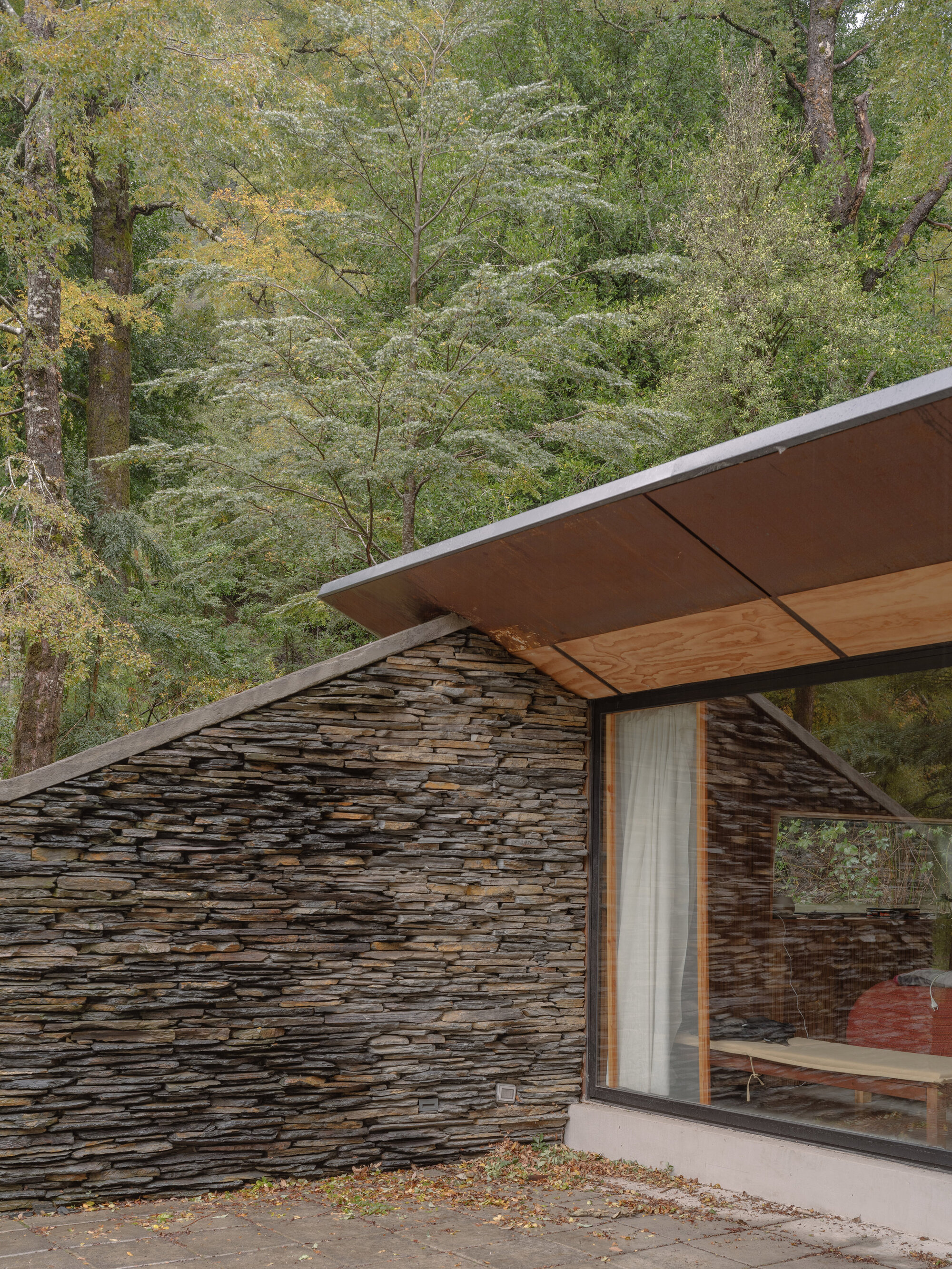 Corner junction of layered stone wall with timber-lined soffit and glazing overlooking the wooded hillside