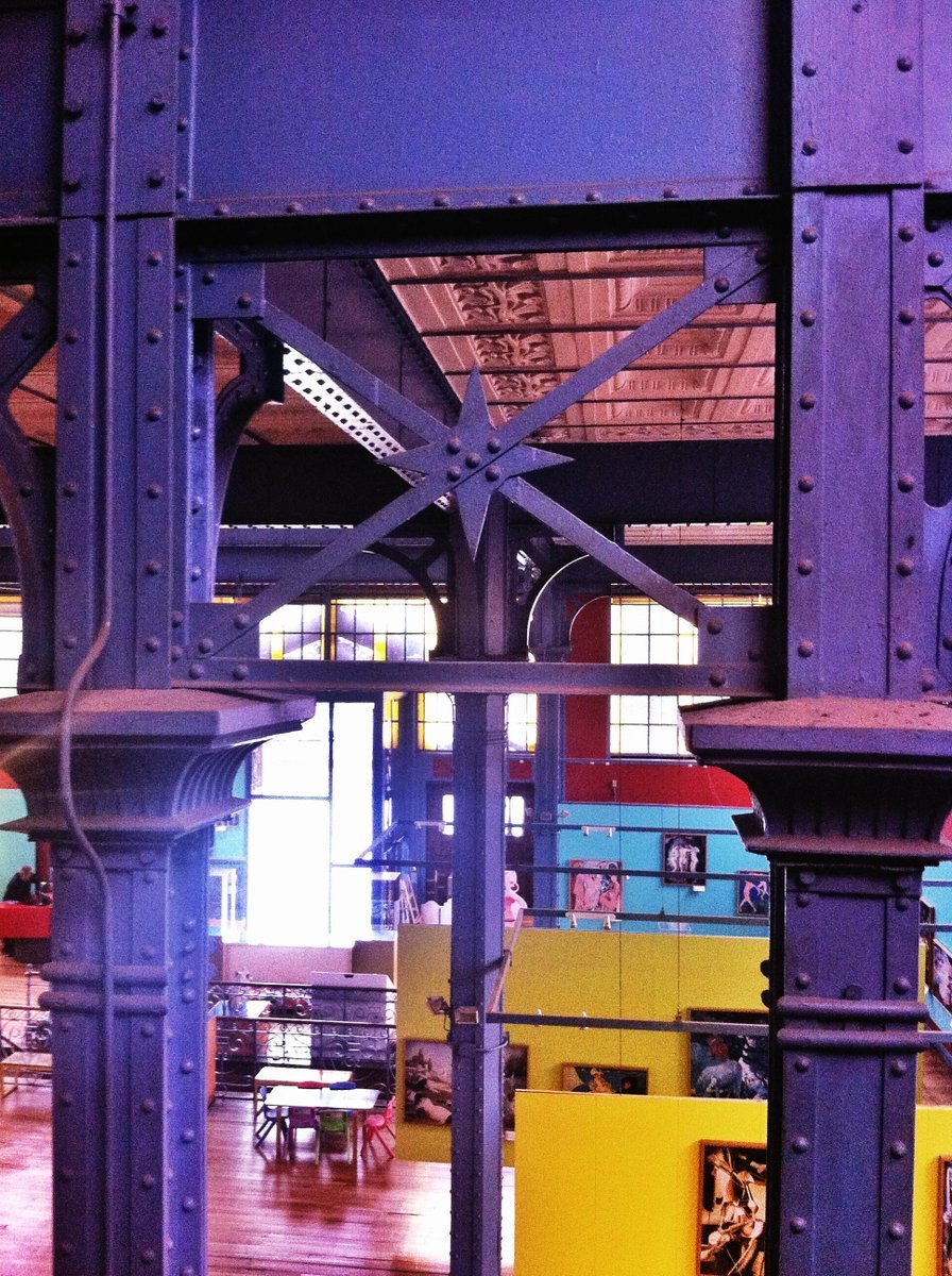 View through the riveted steel trusses at dusk with industrial ceiling and colored exhibition spaces visible beyond