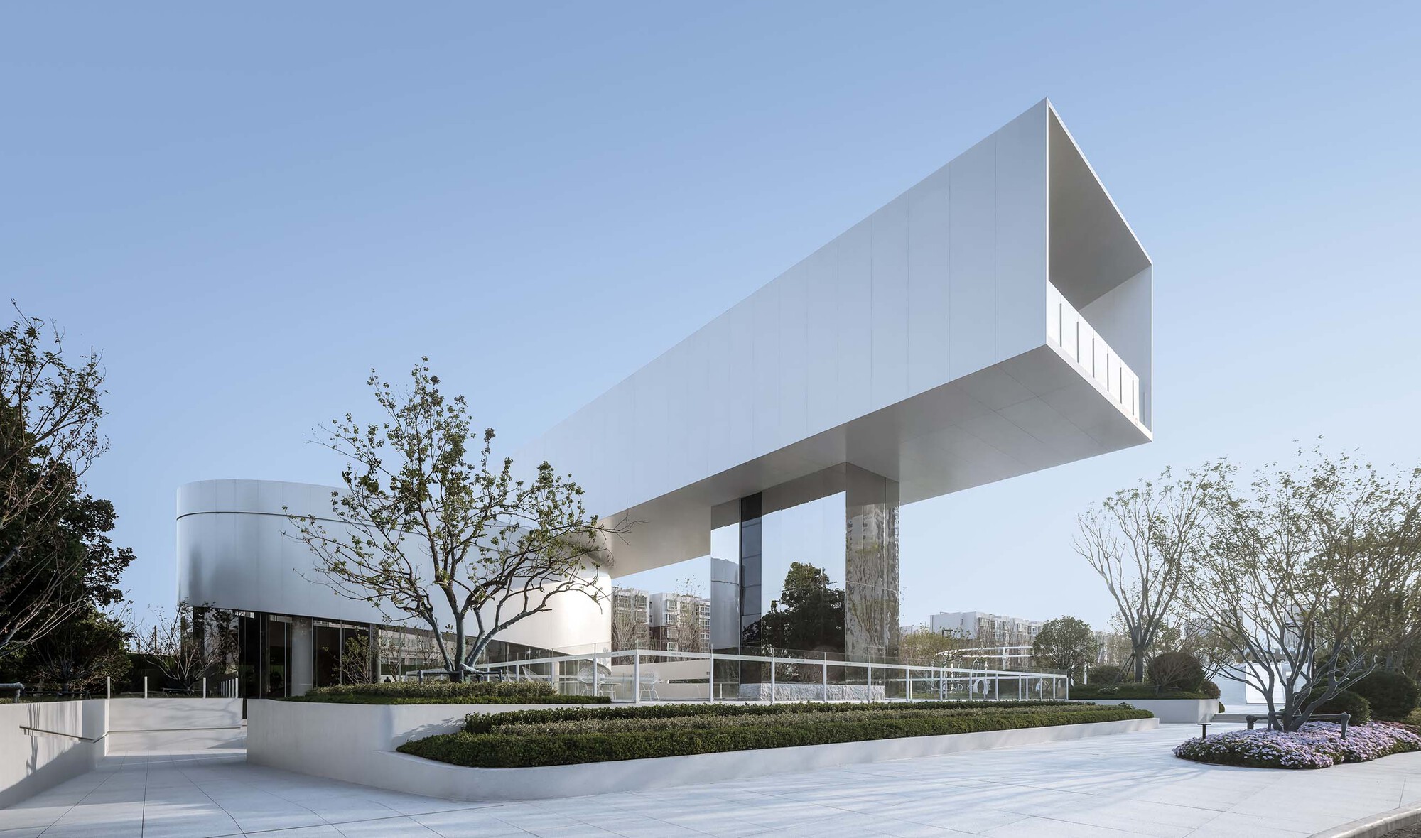 White cantilevered volume raised on columns above a paved plaza with trees and planted beds