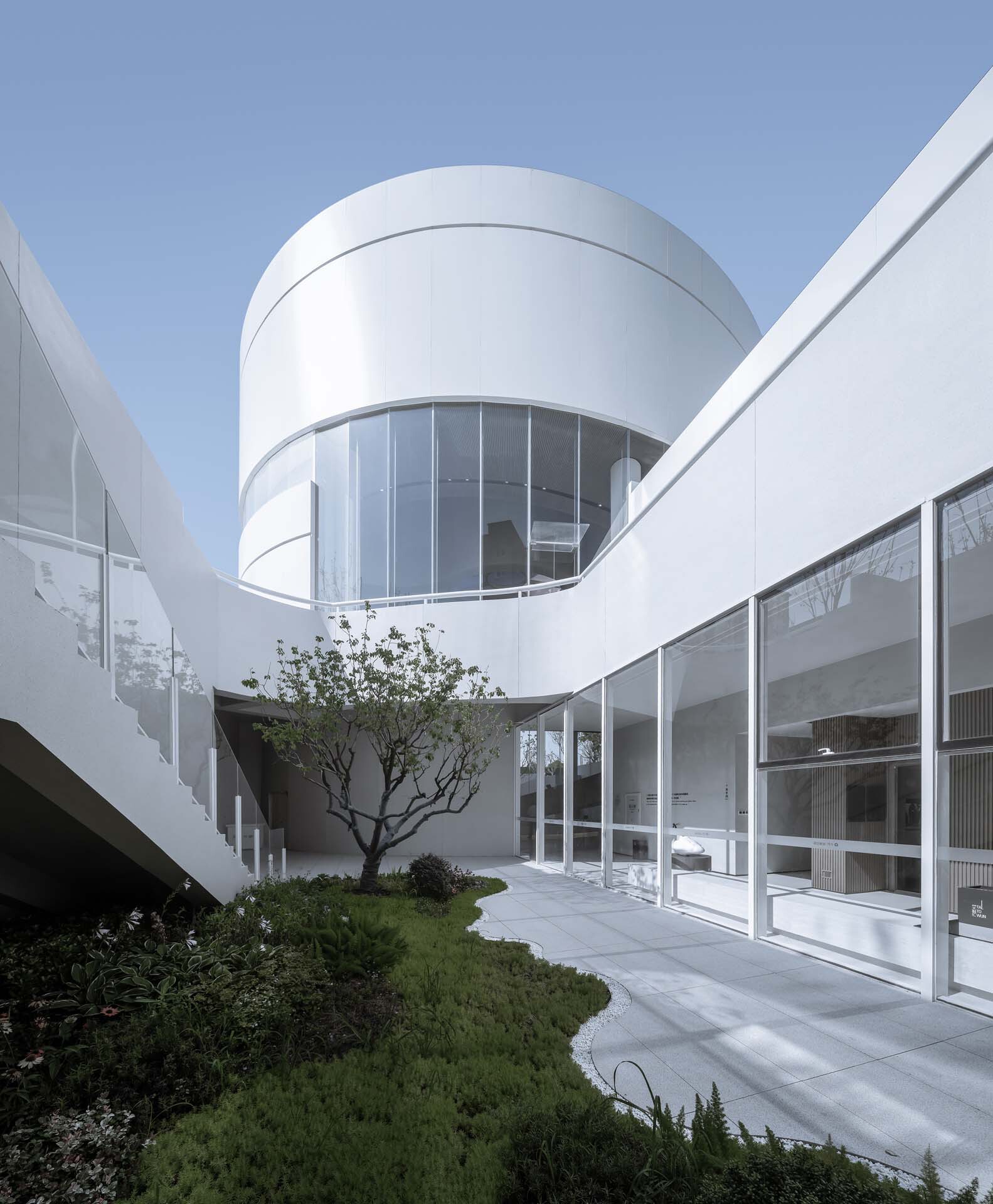 Courtyard with cylindrical glazed volume and planted beds beneath a clear sky