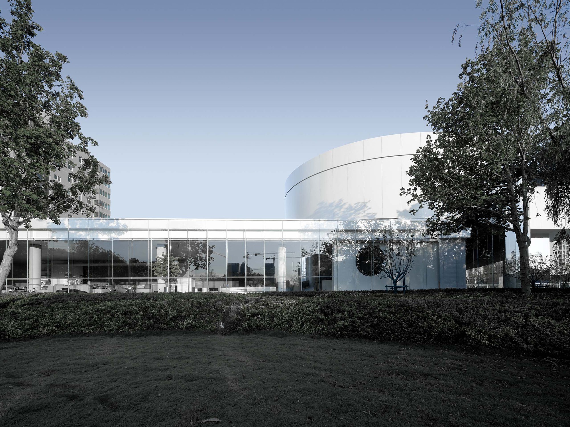 Glass-walled pavilion connecting to cylindrical volume with mature trees framing the composition