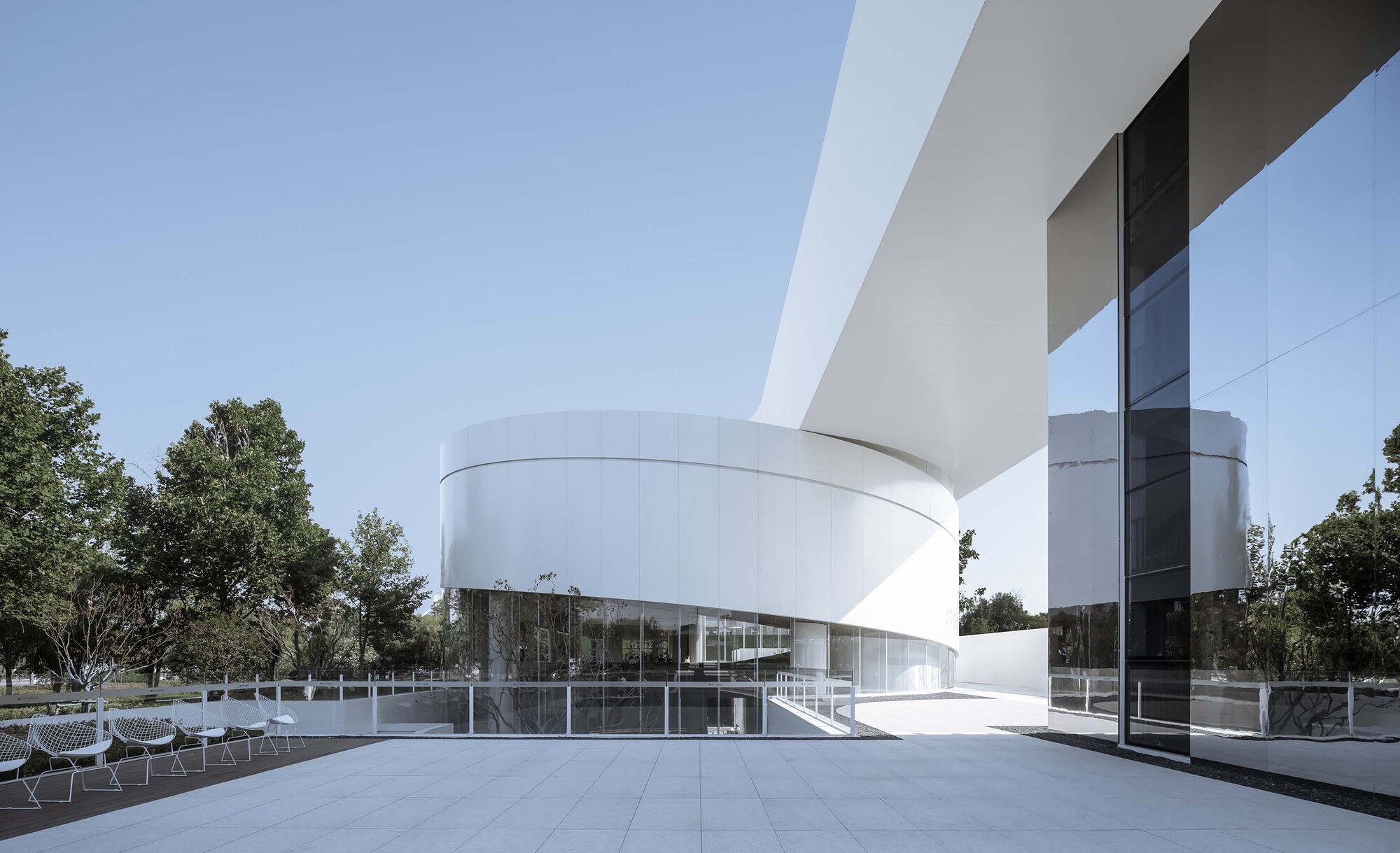 White cantilevered bridge connecting cylindrical volume to glass pavilion with pool and trees beyond