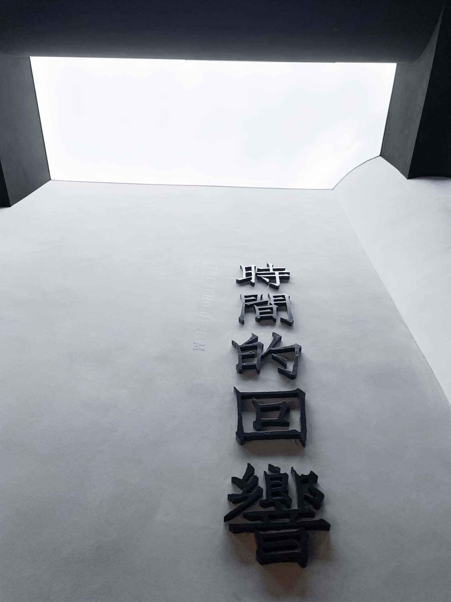 White interior wall with vertical bronze Chinese characters below a recessed ceiling skylight