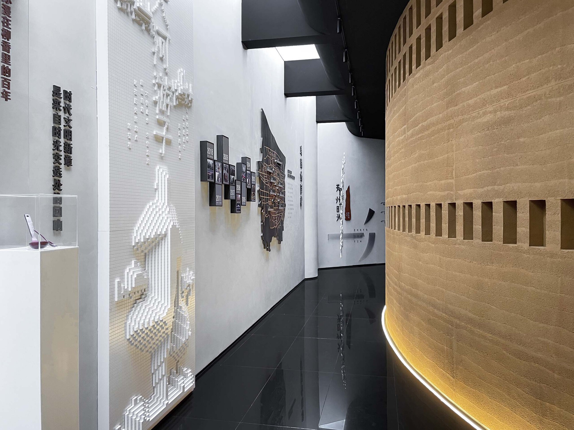 Gallery corridor with white walls featuring relief maps and exhibits alongside a curved timber-clad volume with uplighting