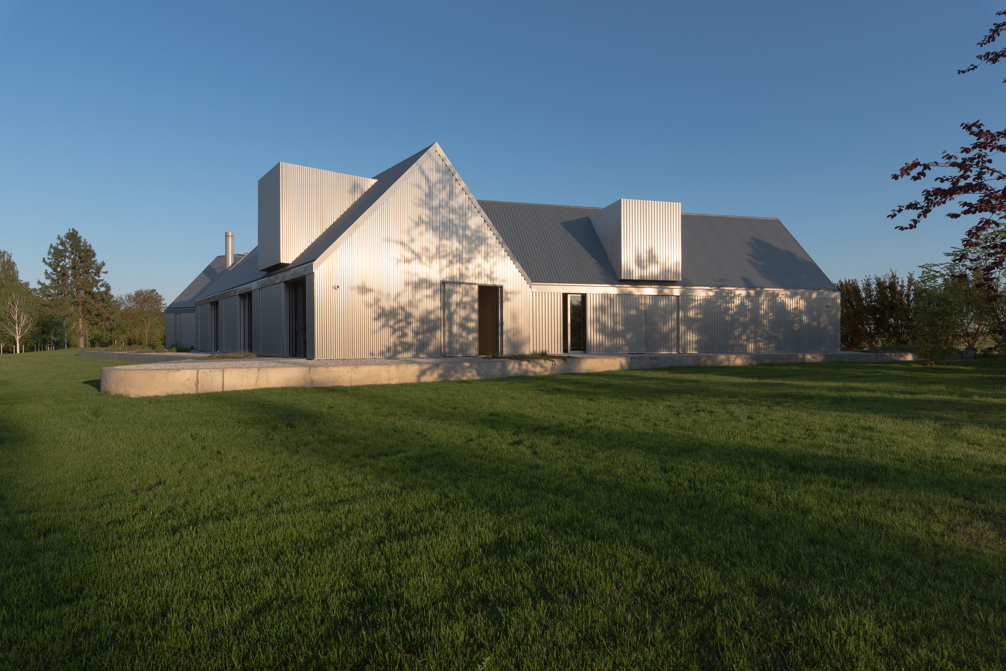 Gabled house with corrugated metal cladding and stone plinth set in a green lawn at dusk