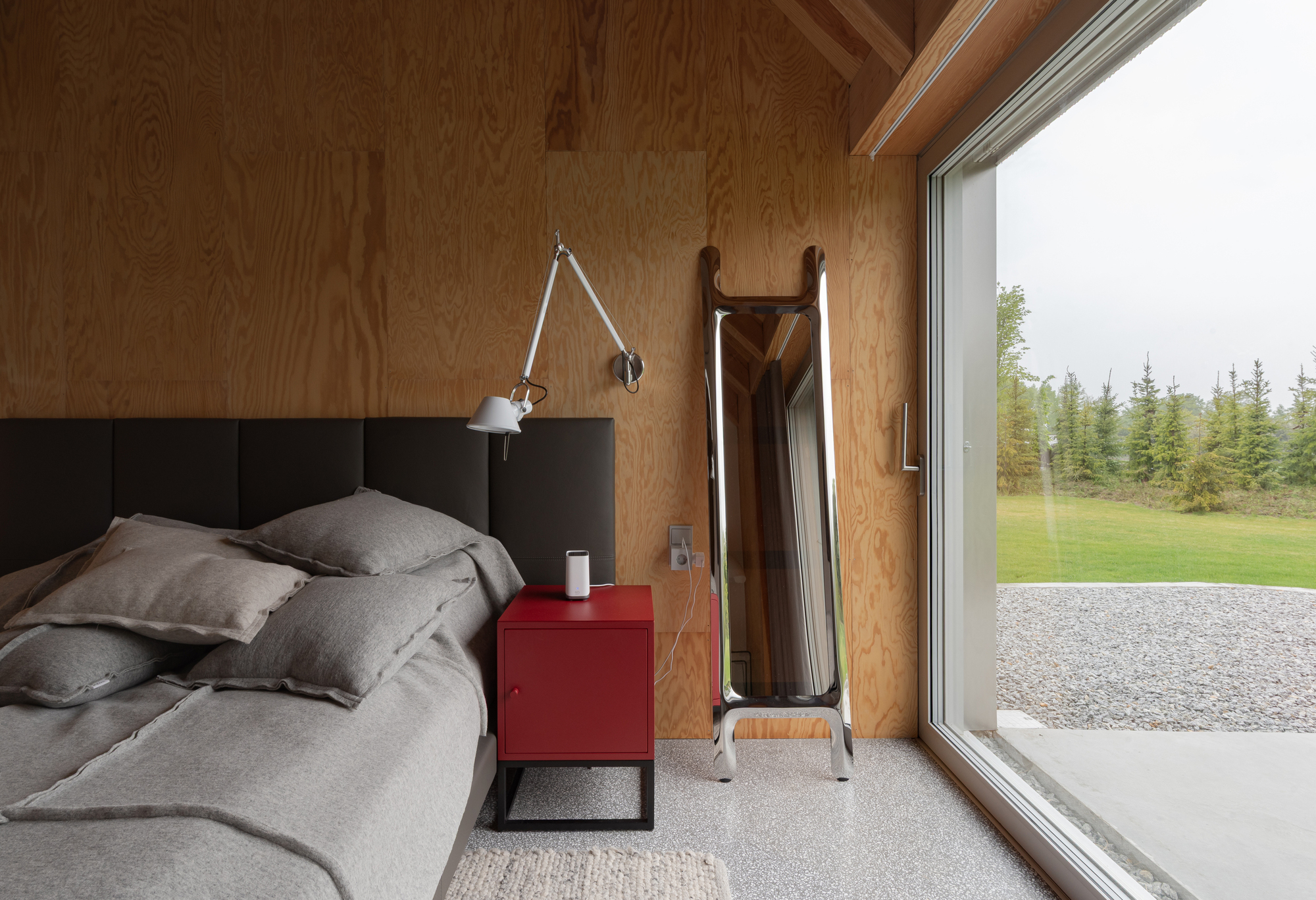 Bedroom with plywood walls and floor-to-ceiling glazing overlooking a gravel path and green field