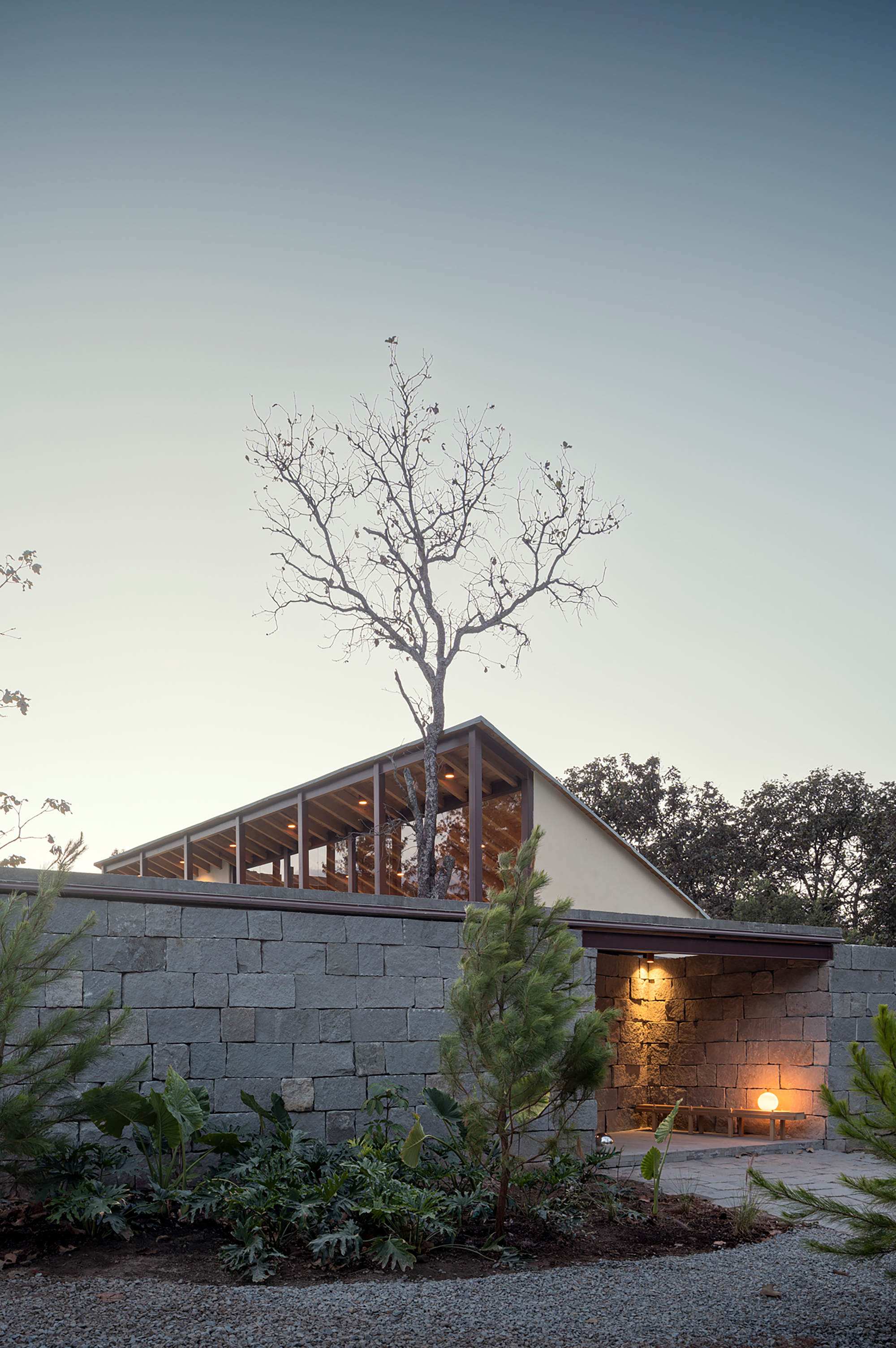 Stone block wall with exposed timber roof structure and bare tree silhouette at dusk