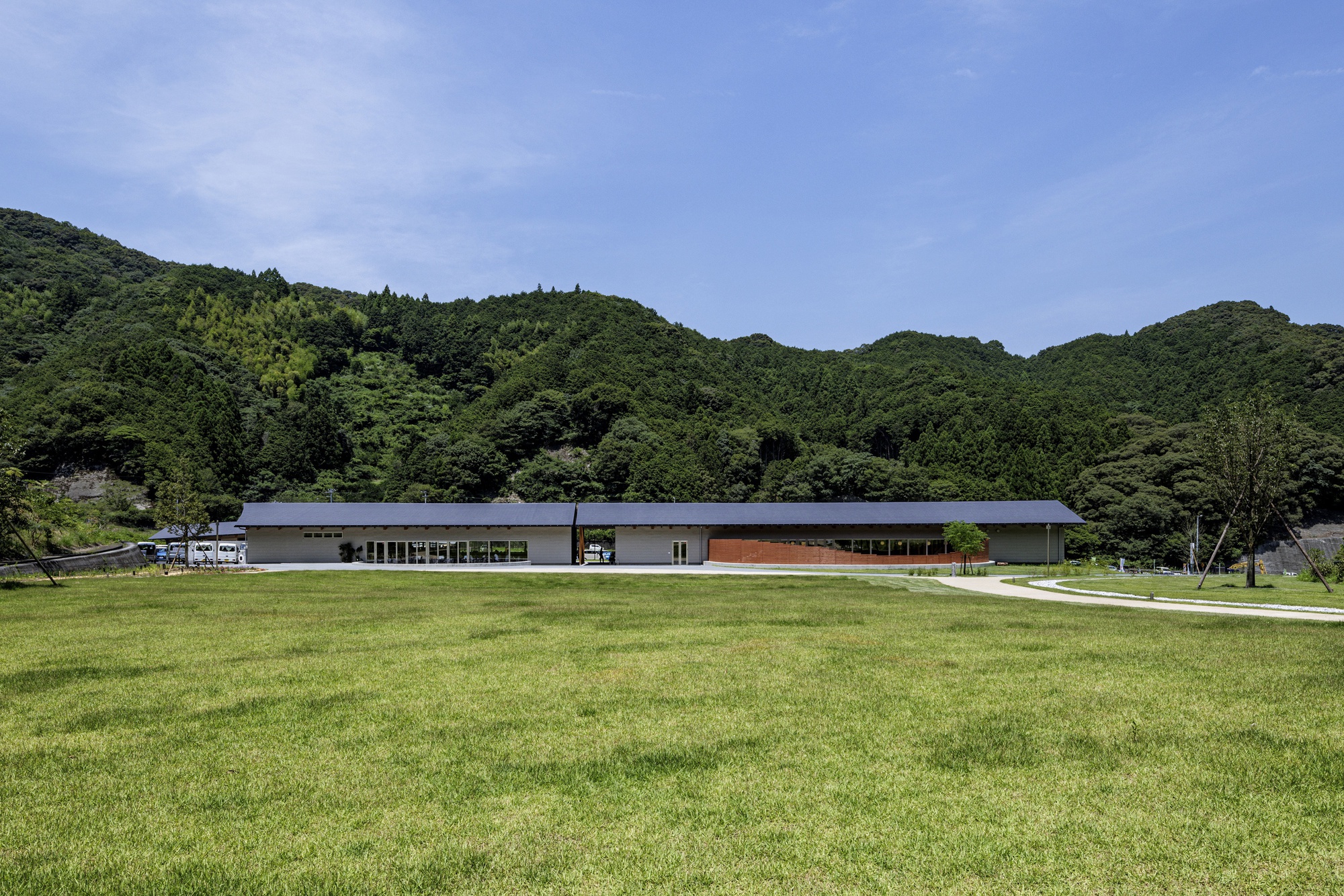Long horizontal building with dark roof and timber cladding across an open grass field backed by forested hills