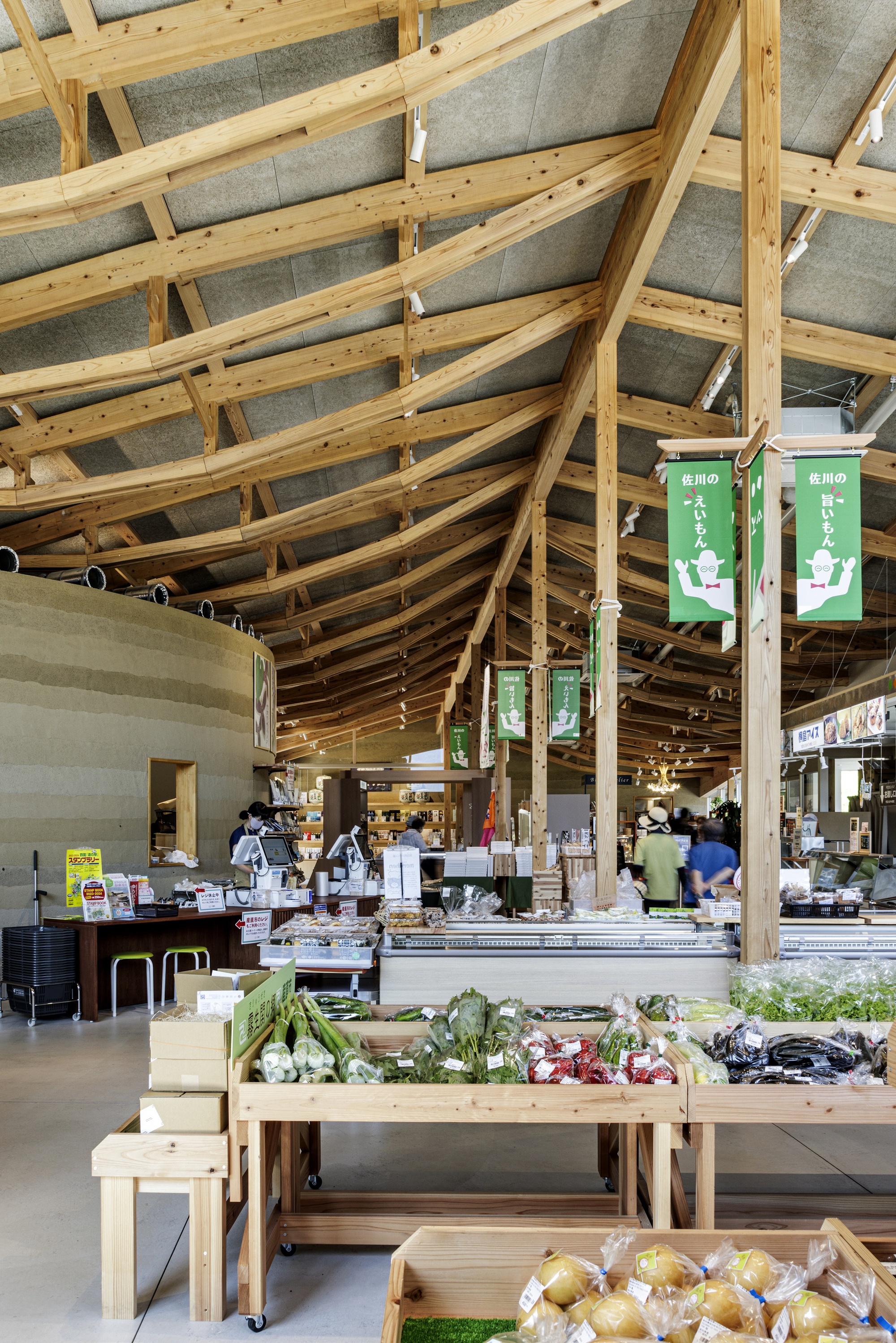 Produce market interior with timber display tables beneath curved timber beams and suspended green banners