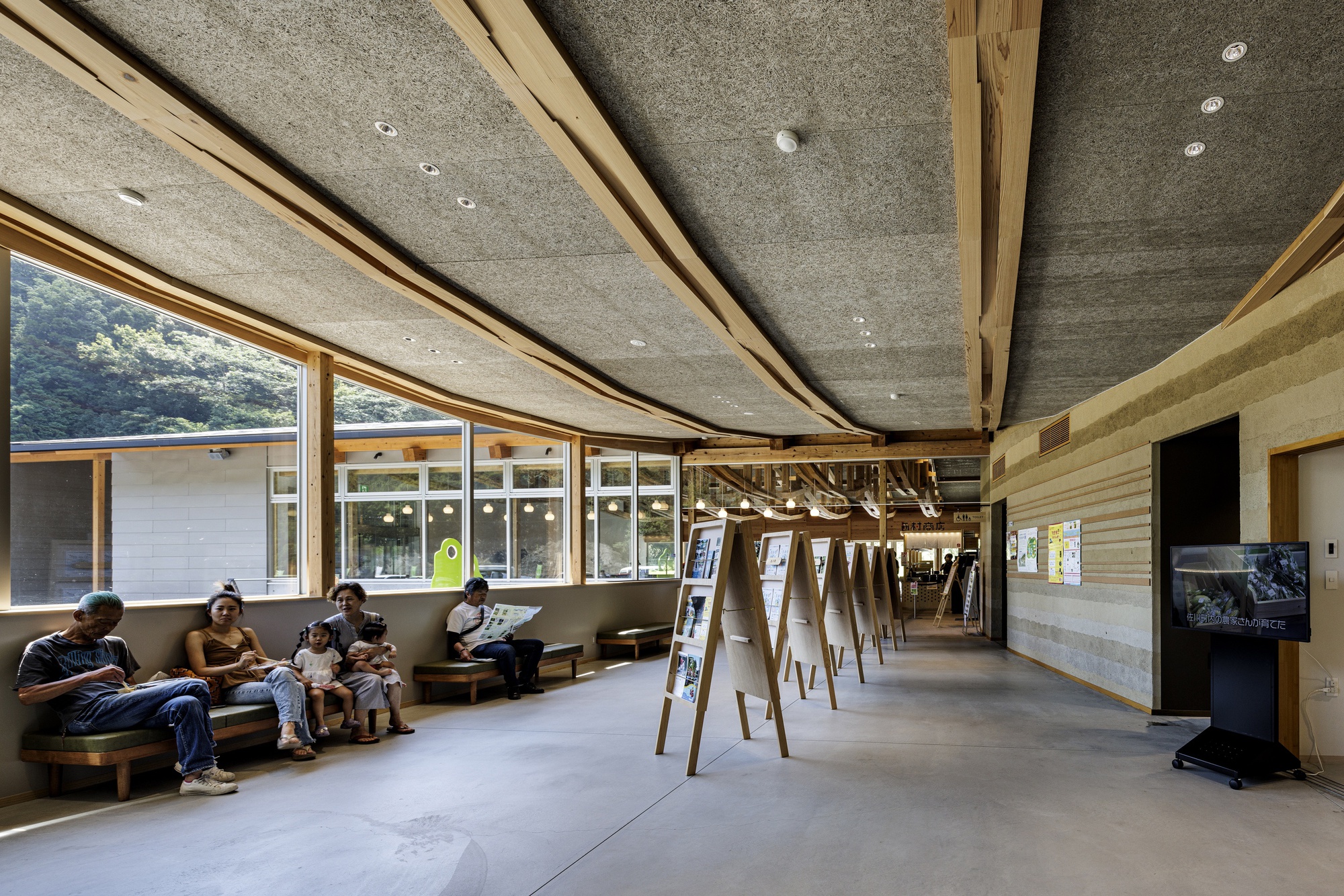 Interior corridor with angled timber ceiling beams, concrete panels, and visitors seated near windows overlooking green hillside