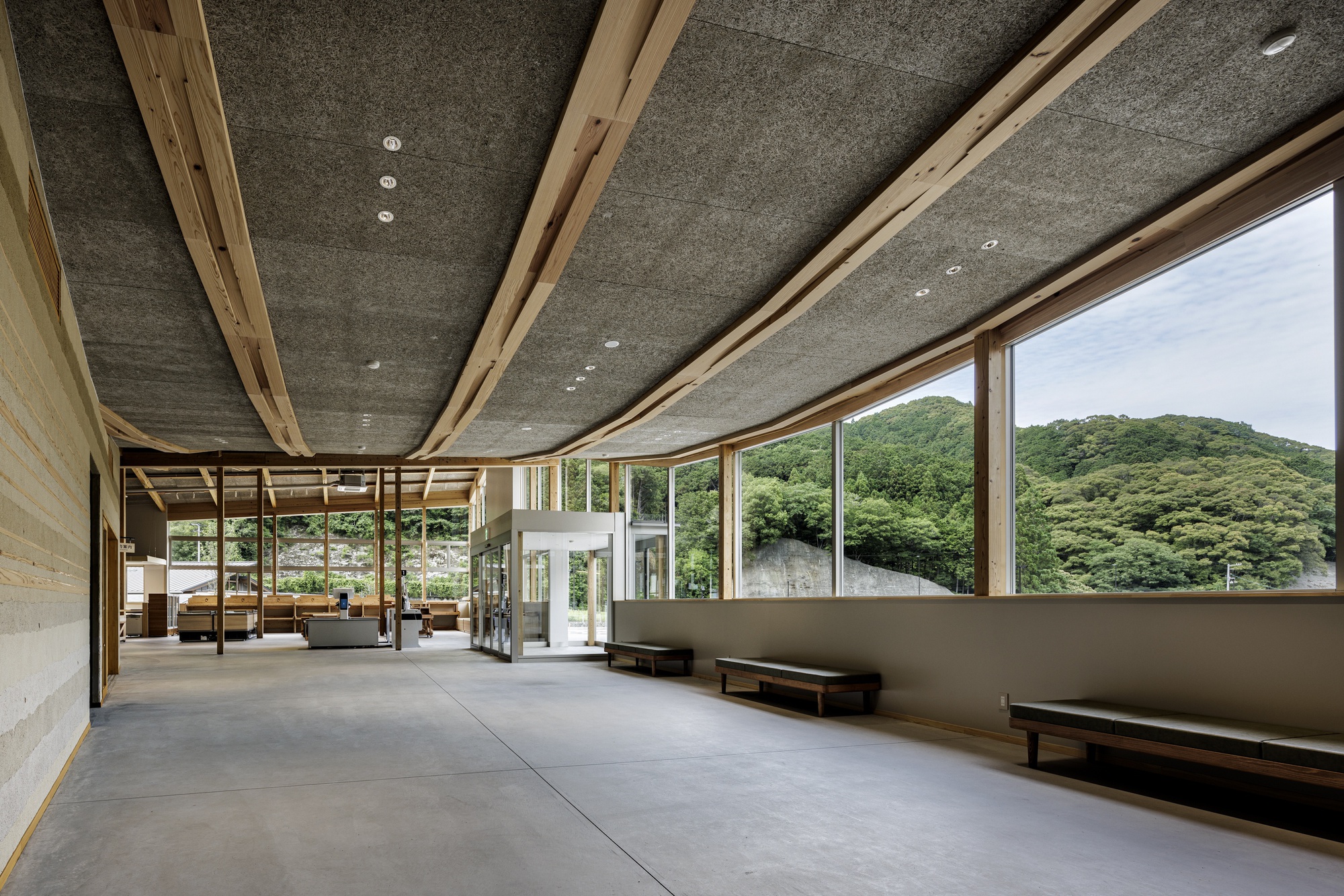 Long interior hall with exposed timber roof structure, concrete ceiling panels, and continuous ribbon windows framing forested mountains