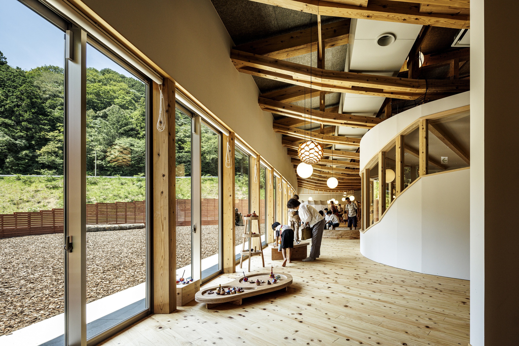 Curved white wall alongside glazed corridor with timber posts, exposed wood ceiling beams, and visitors browsing displayed objects