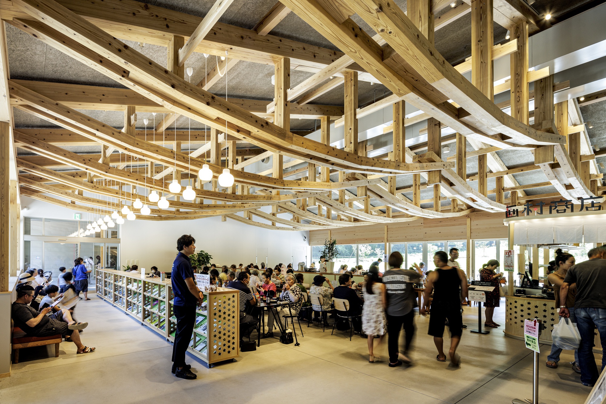 Busy interior space with layered timber truss ceiling, pendant lighting, and visitors gathered around low display cases