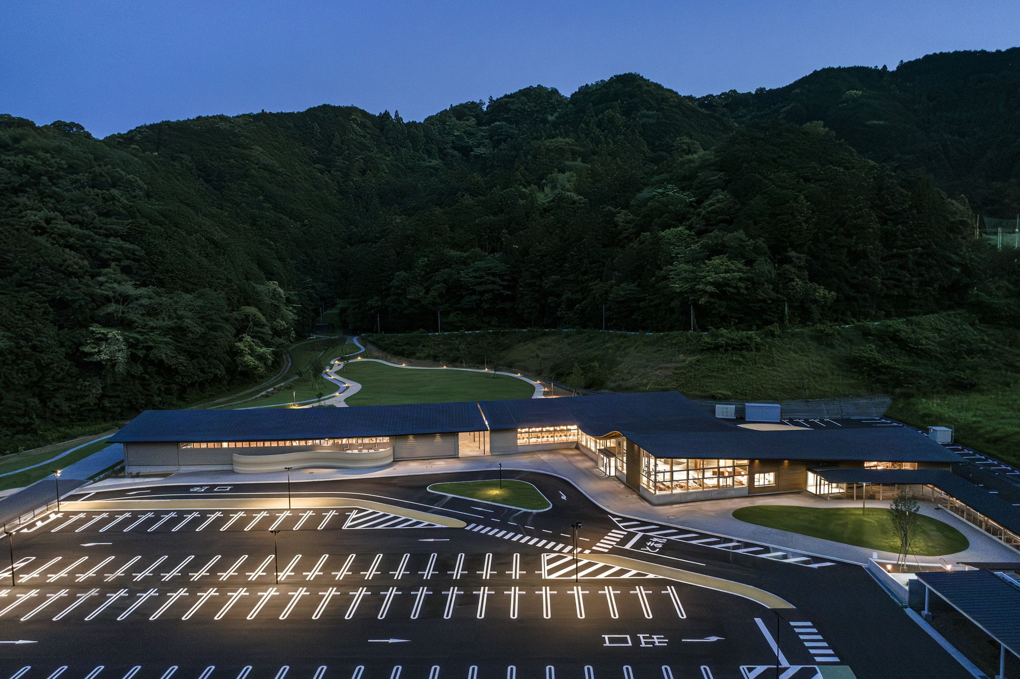 Illuminated glass facade and parking lot at twilight with forested hills rising behind