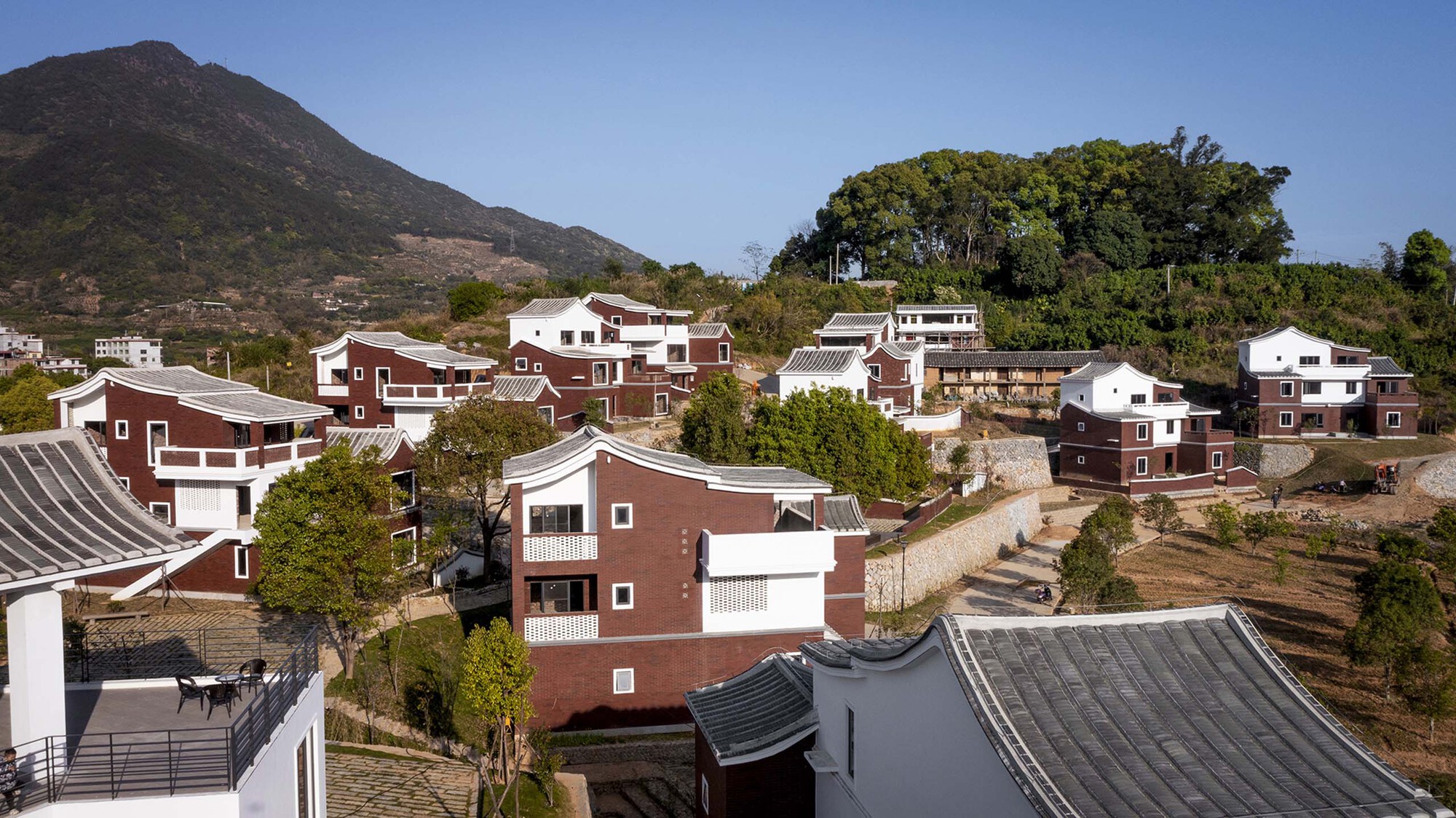 Housing cluster on sloped terrain with red brick and white volumes under curved tile roofs with mountains behind