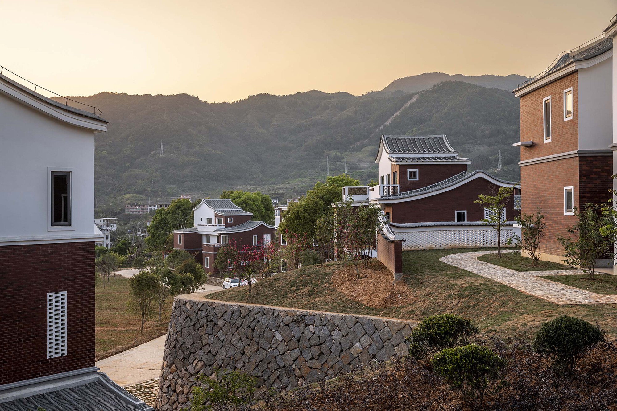 Stone retaining wall and planted slope with housing volumes scattered across the hillside at dusk