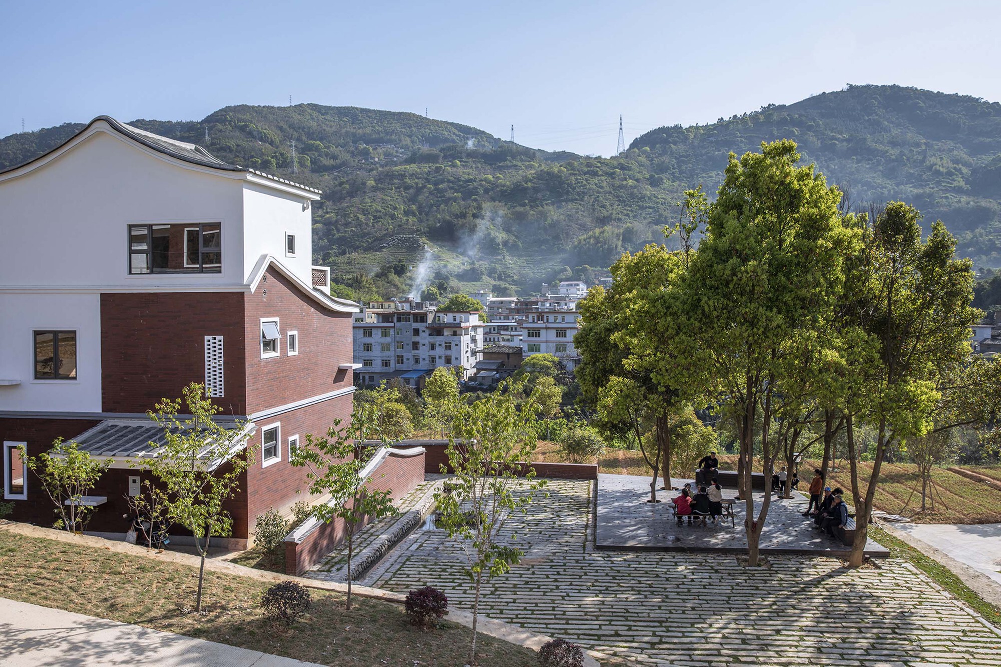 Tree-lined community plaza with residents gathering beneath a green mountain range