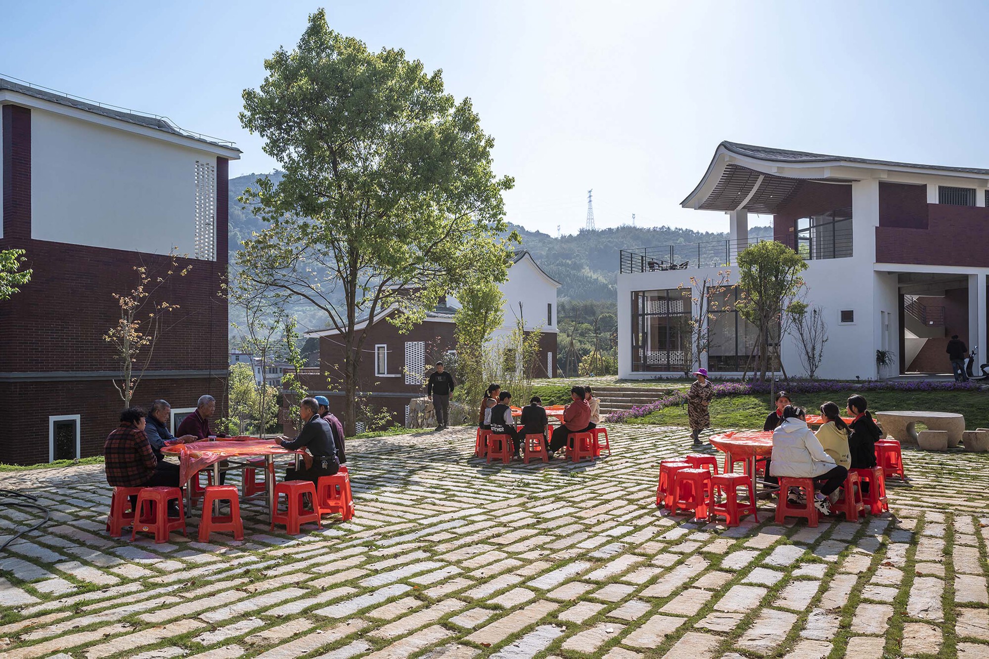 Residents seated at red tables and stools on a permeable paving grid in midday sun