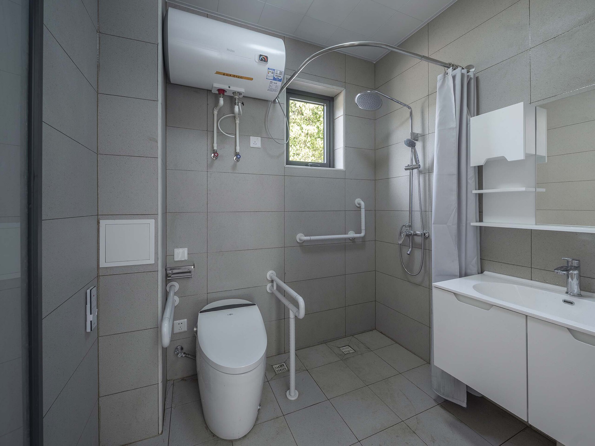 Accessible bathroom with grey tile walls, grab bars, and a small window above the shower