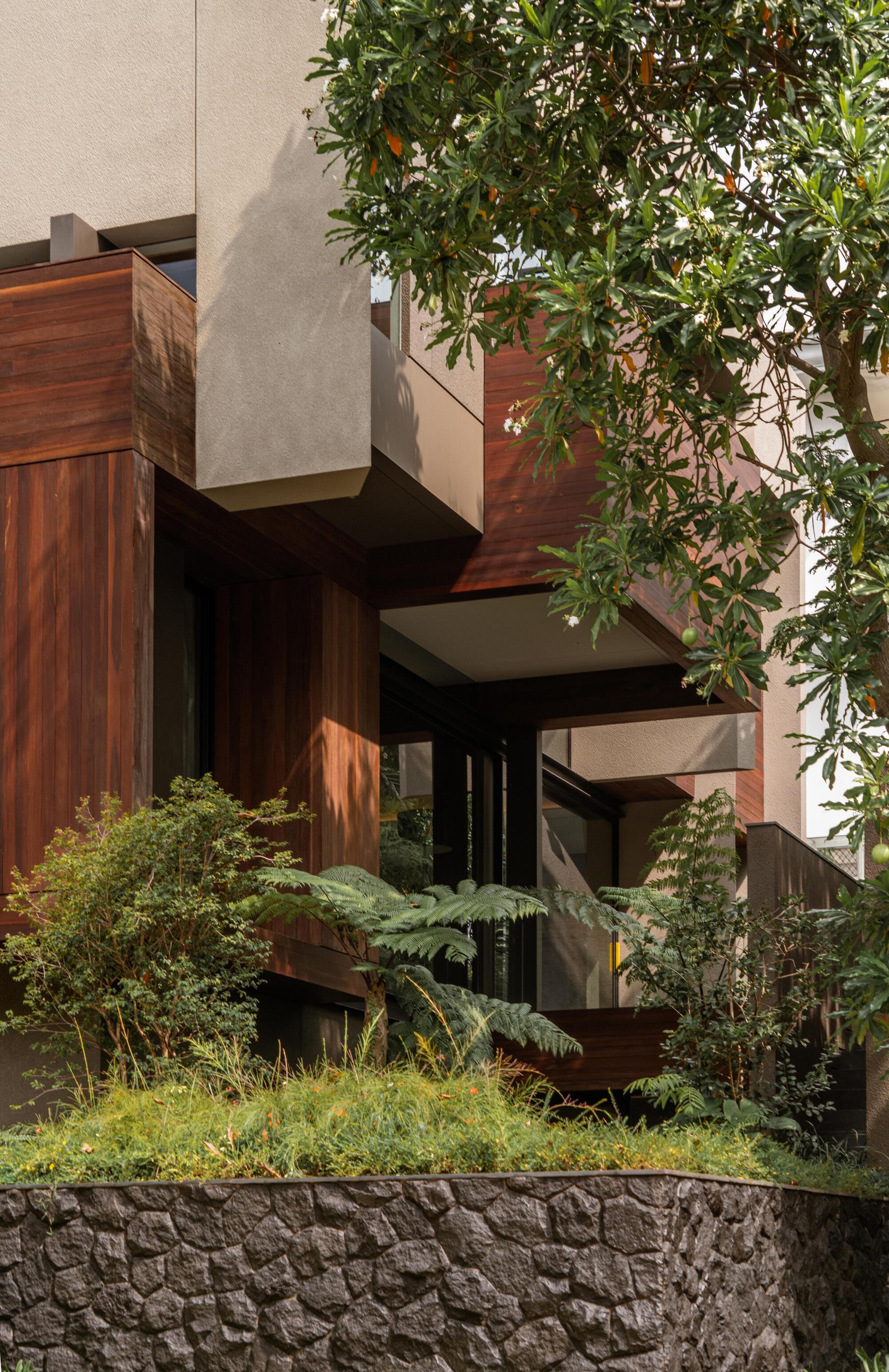 Street view of stacked timber-clad volumes with planted terraces framed by trees and stone retaining wall