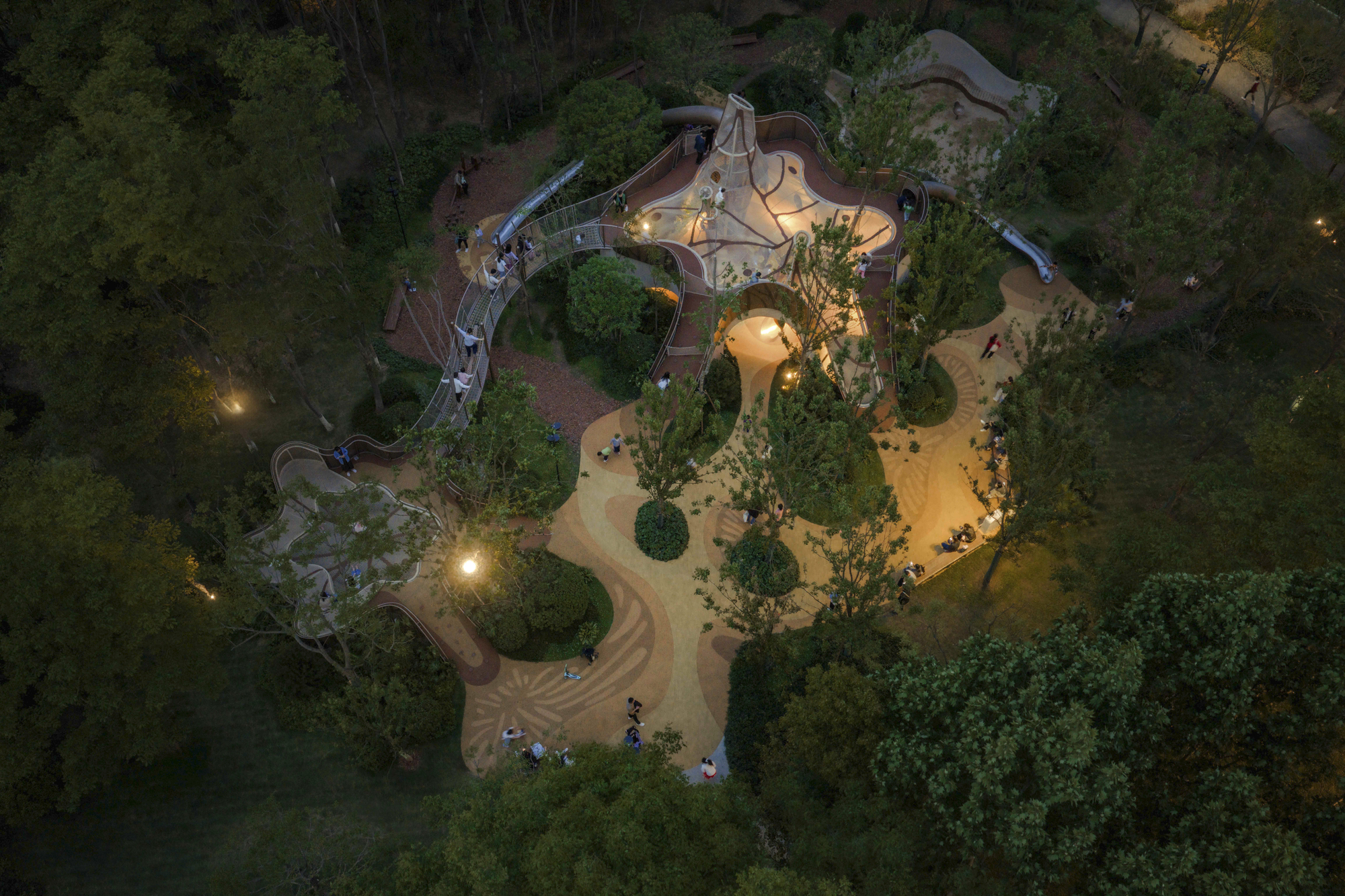 Aerial view at dusk of a curving playground structure with sand paths winding through trees