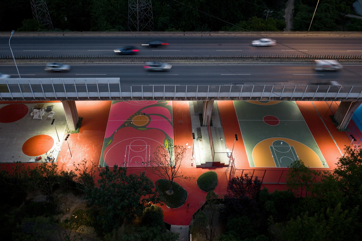 Illuminated basketball courts in pink and orange beneath a highway with evening traffic overhead