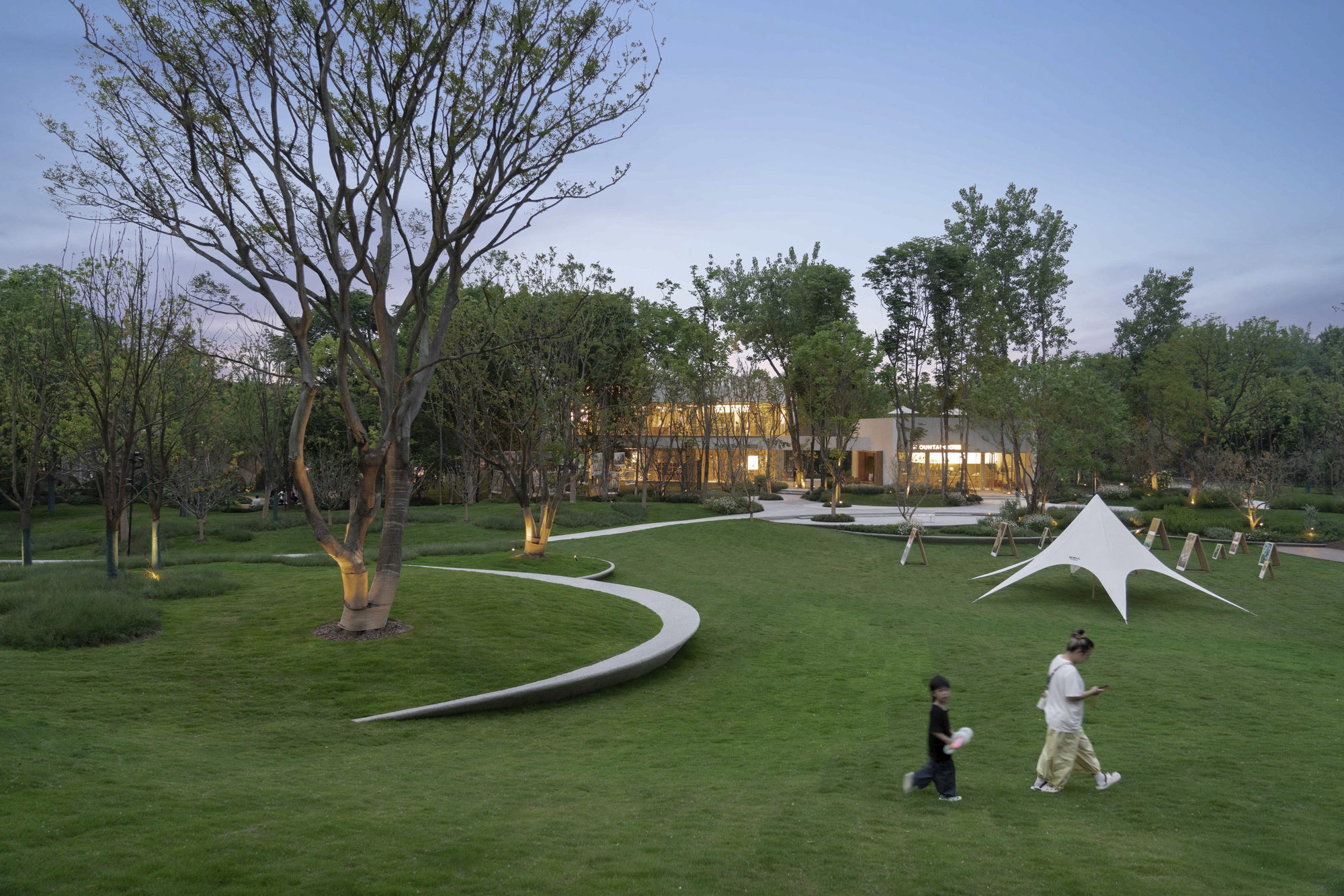Curved pathways leading through lawn toward illuminated building at dusk with visitors strolling