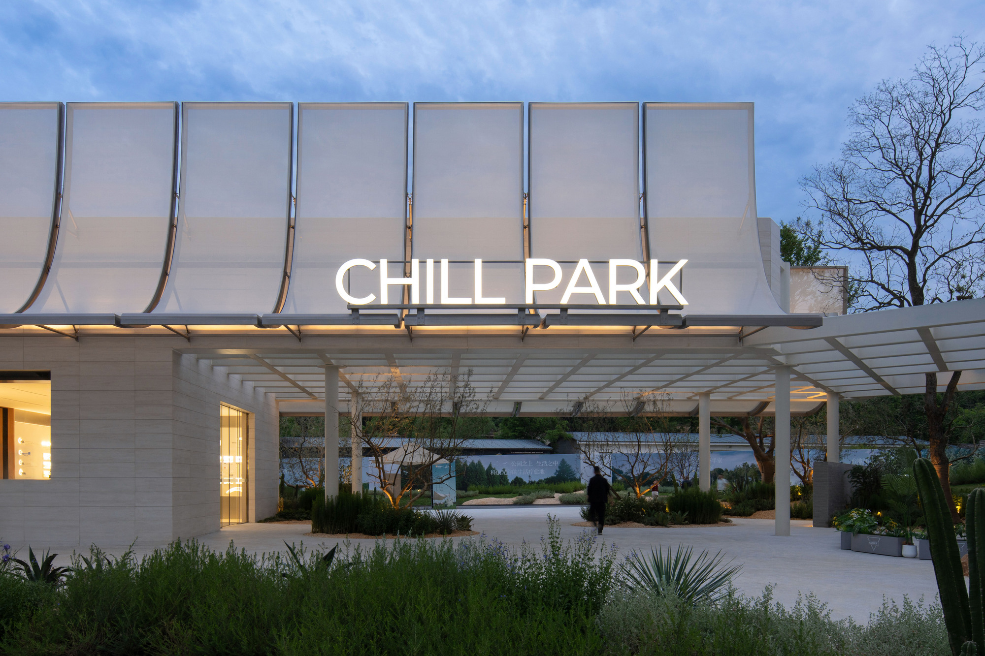 Illuminated entrance canopy with curved fabric roof panels and white pergola structure at dusk