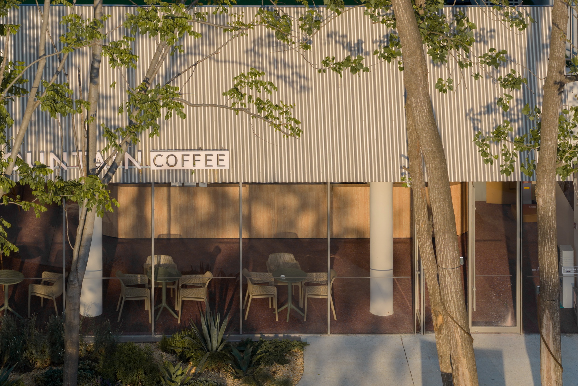 Corrugated metal wall with ribbon windows casting dappled tree shadows at golden hour