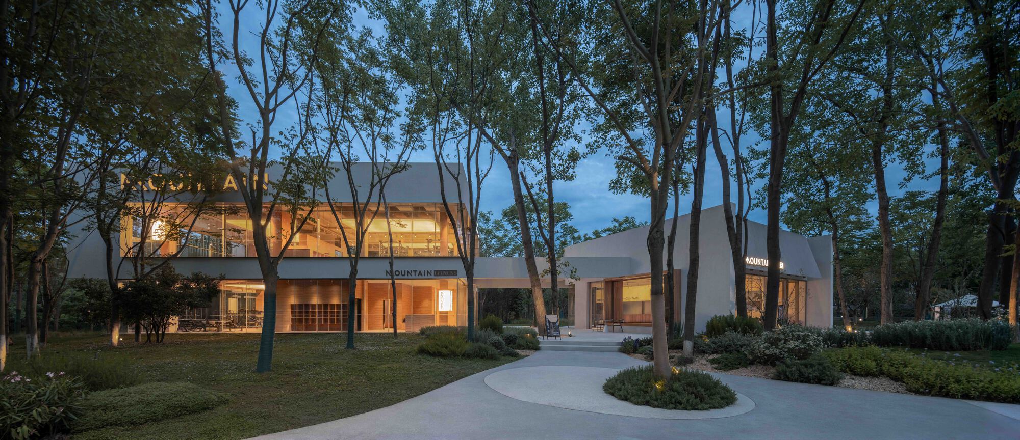 Illuminated volumes connected by a covered pathway through a wooded site at dusk