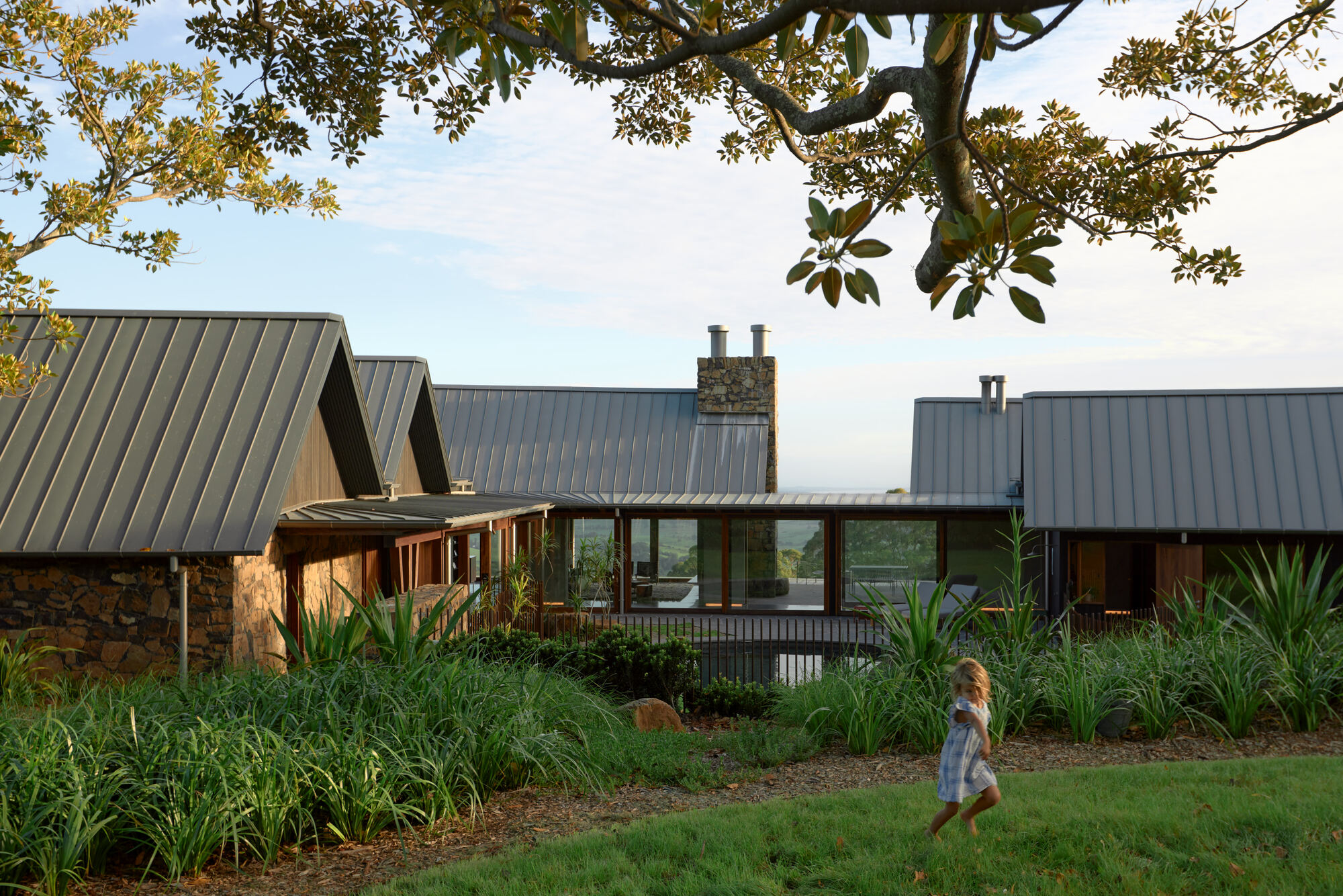 Garden view of linked gable volumes with metal roofing and stone walls as a child runs across the lawn