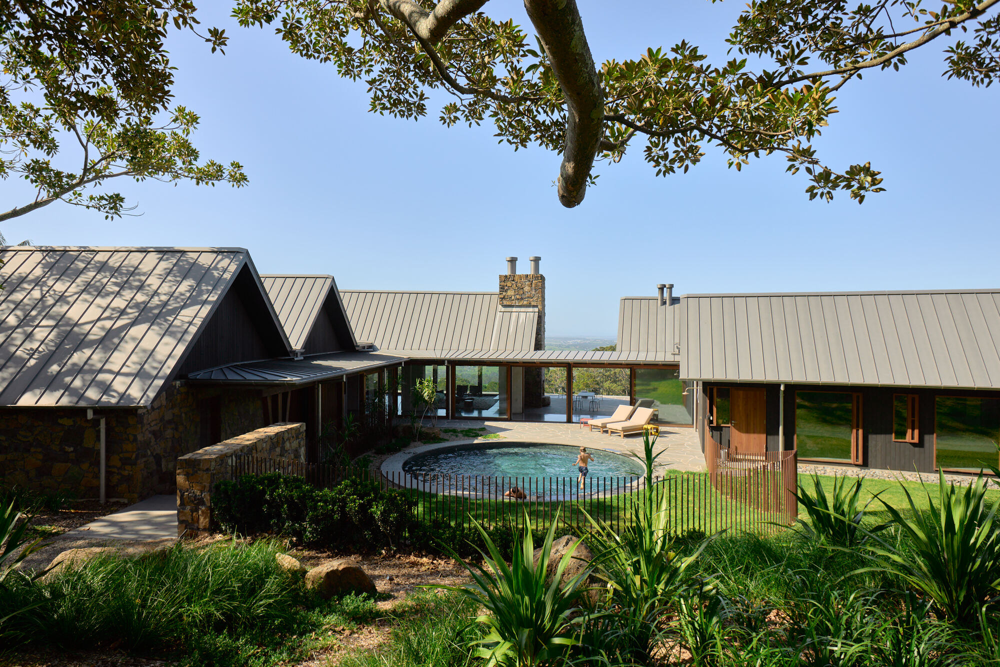 Courtyard with circular pool framed by pitched roofs and native plantings beneath a spreading tree